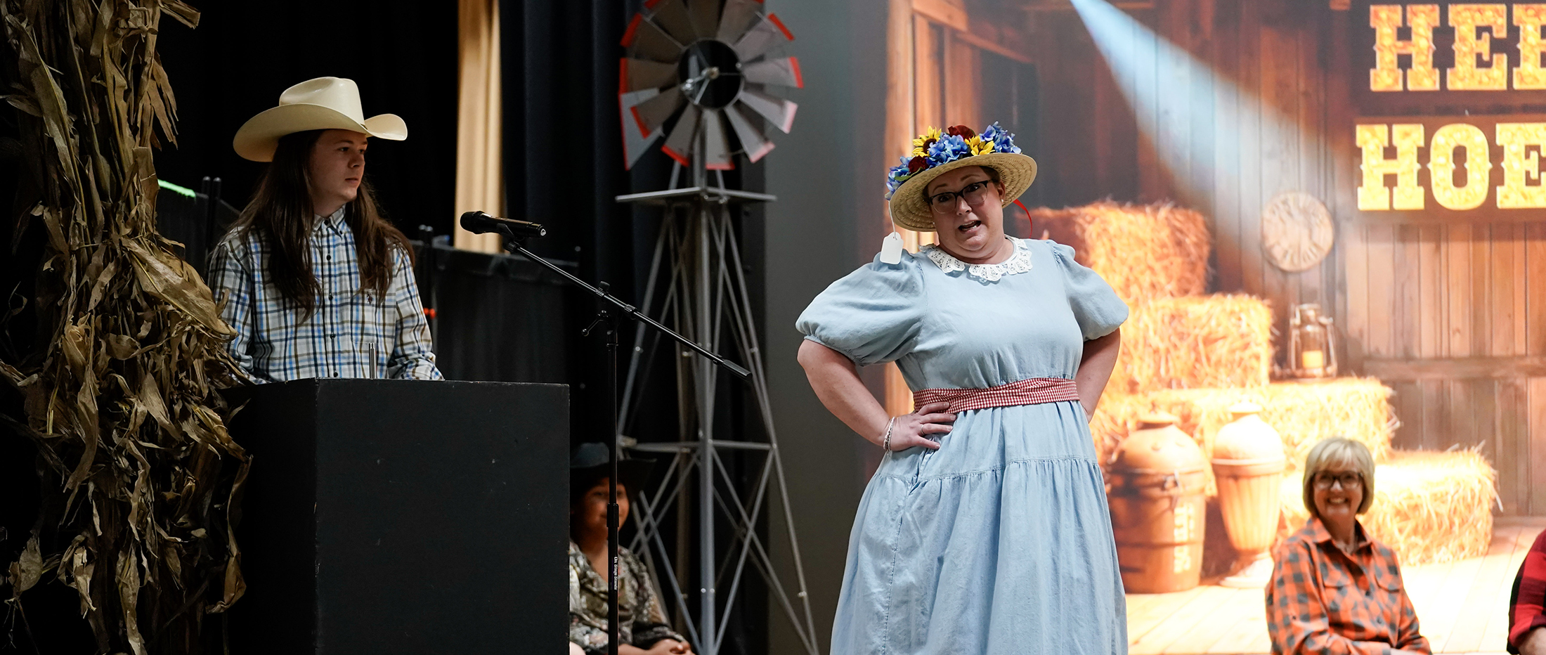 young man in cowboy hat behind podium with woman dressed as Minnie Pearl with hands on hips