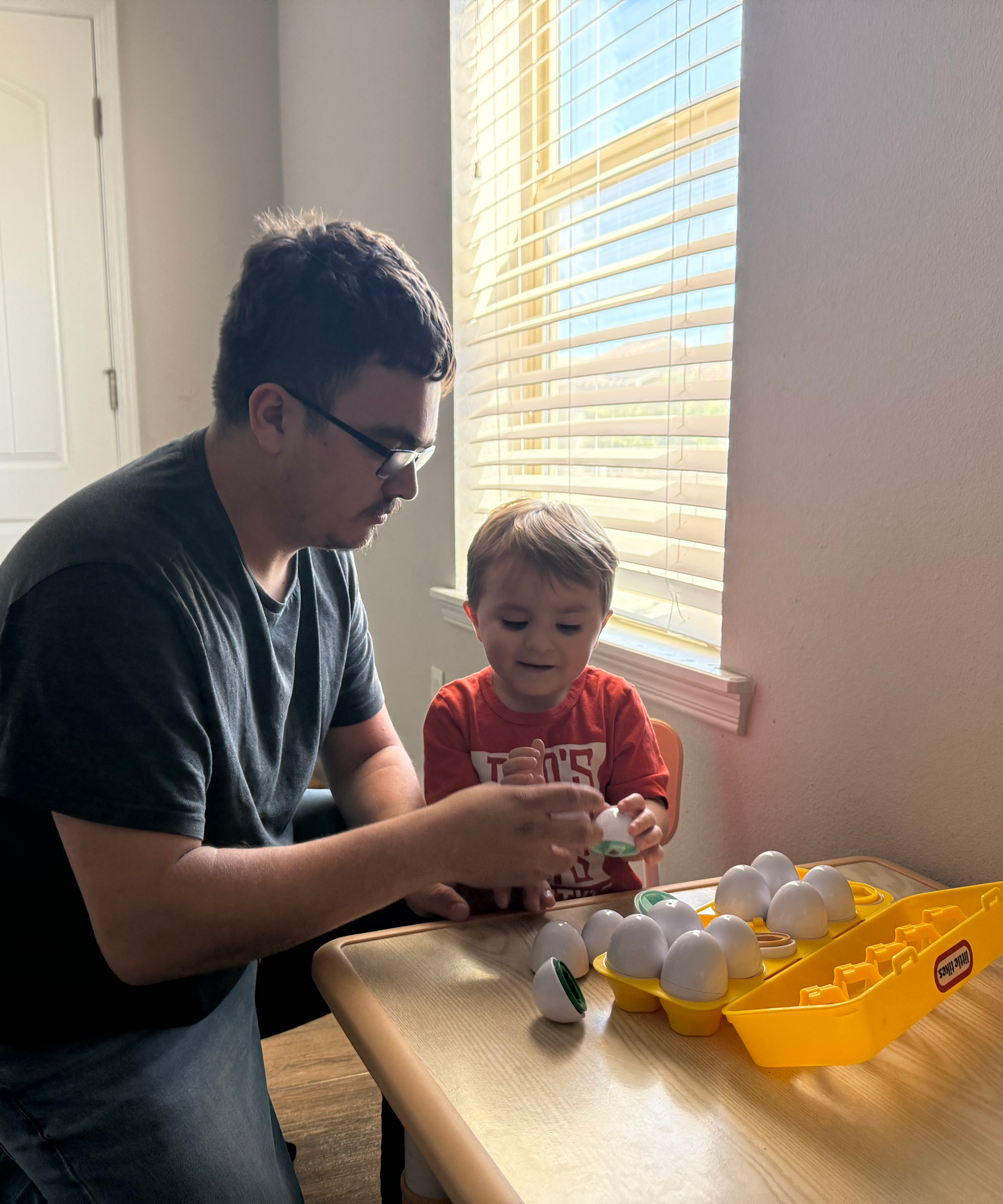 A man and a young boy are playing with toy eggs at a table, with a window and blinds in the background.
