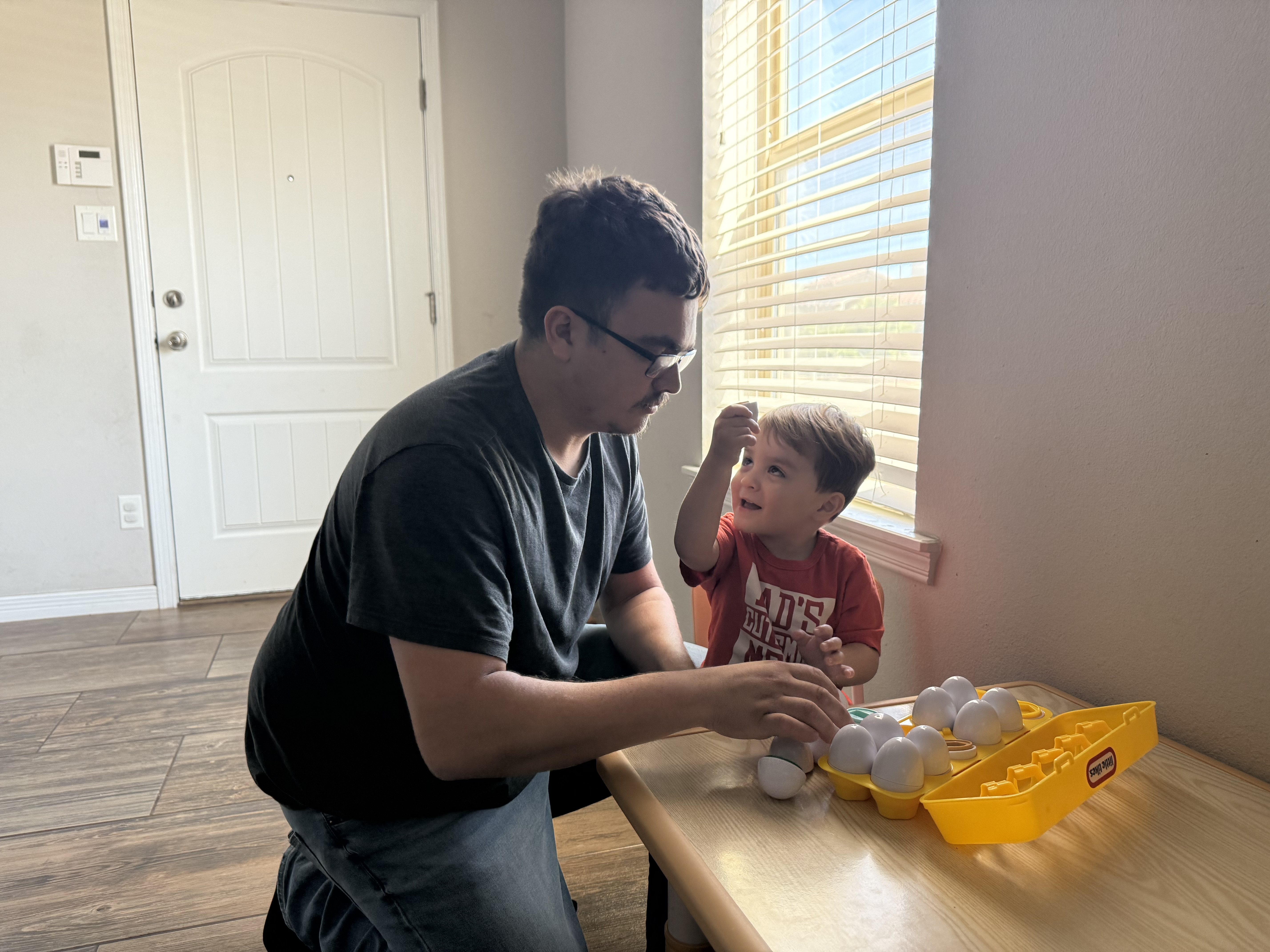 A man and a young boy are playing with toy eggs on a table in the foreground, with a bright window and a door in the background.