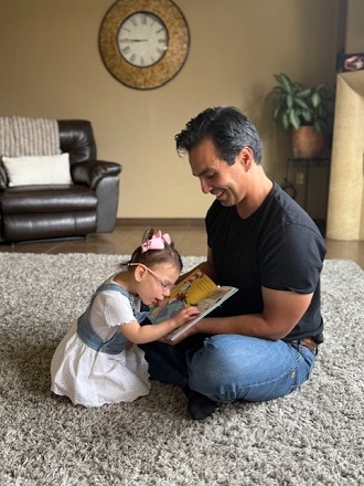 A father and daughter sit on a plush rug reading a book, with a large clock and a leather armchair visible in the background.