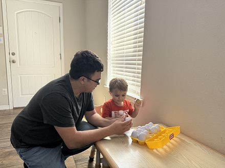 A man and a young child are playing with toy eggs on a table in front of a window, with a door visible in the background.