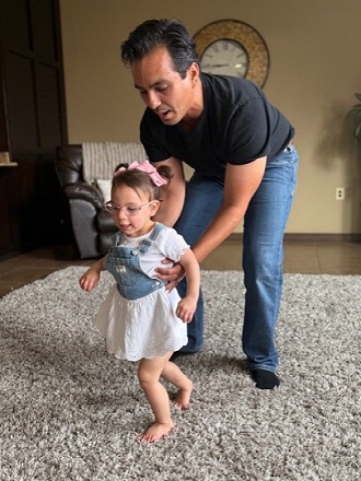 A man in jeans and a black t-shirt helps a young girl in a denim overall dress and pink bow take her first steps on a fluffy rug, with a living room and a clock visible in the background.
