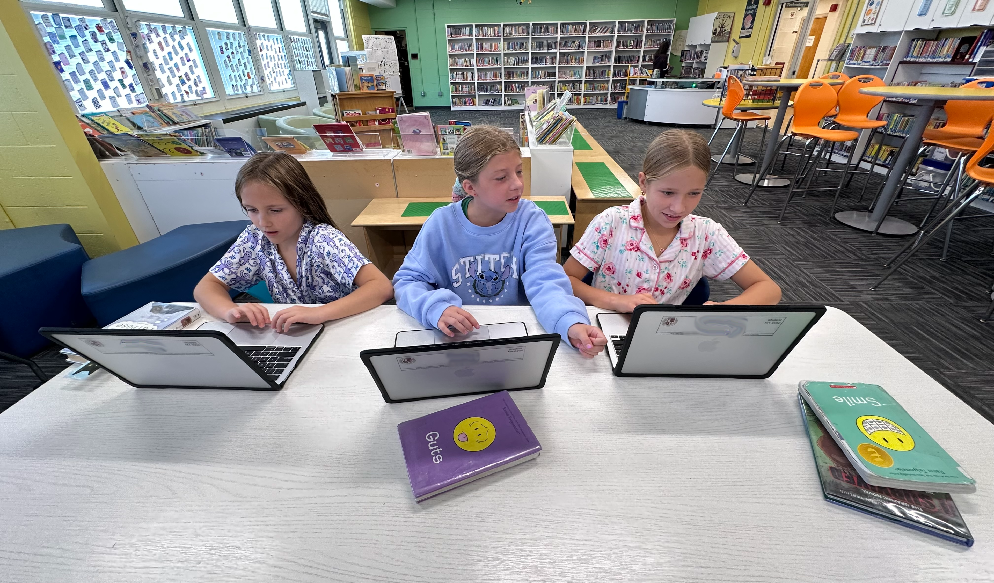 Three young girls focus on their laptops at a table in a brightly lit library with bookshelves and colorful seating in the background.
