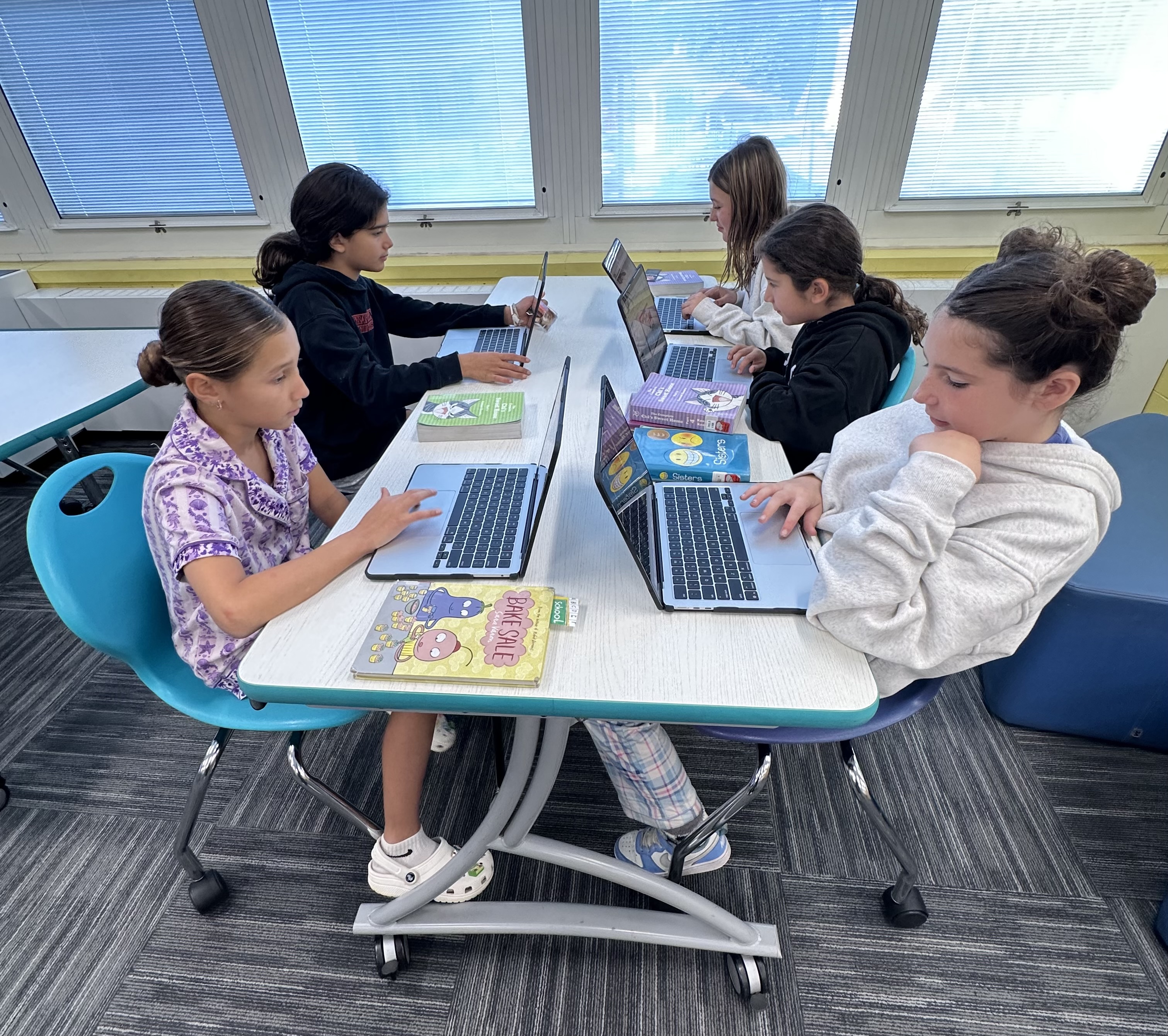 Young girls in the foreground are engaged with laptops at a table, while in the background, more students work at similar stations in a brightly lit classroom with large windows.