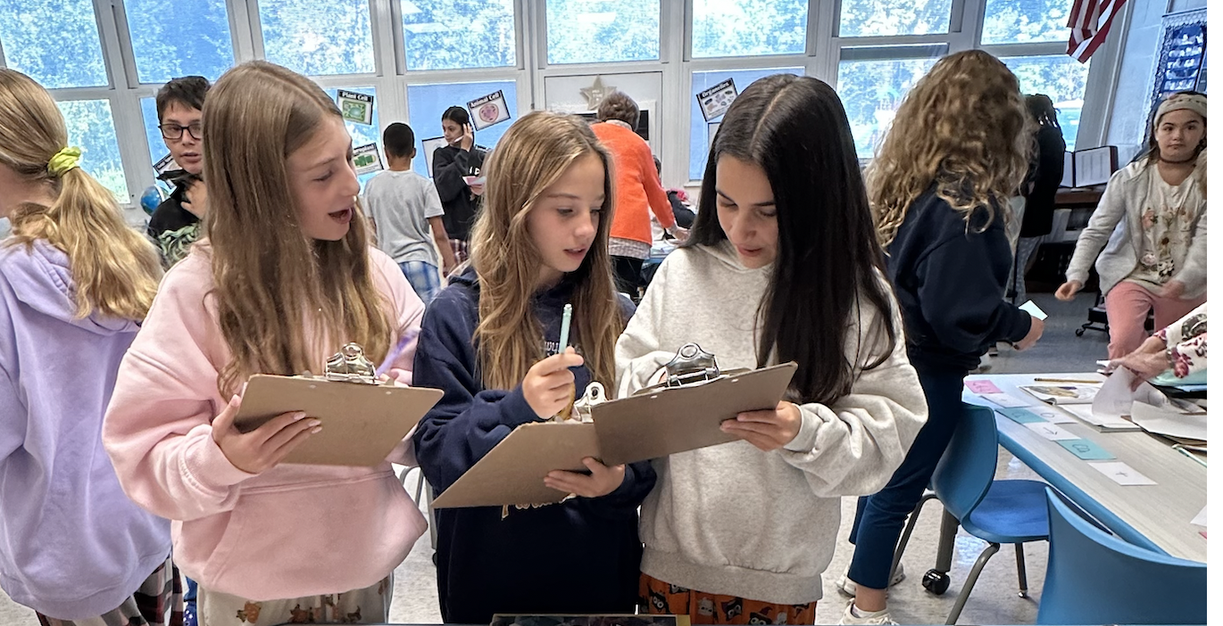 Three young girls in the foreground are intently focused on clipboards, while in the background, other students and adults are engaged in classroom activities.