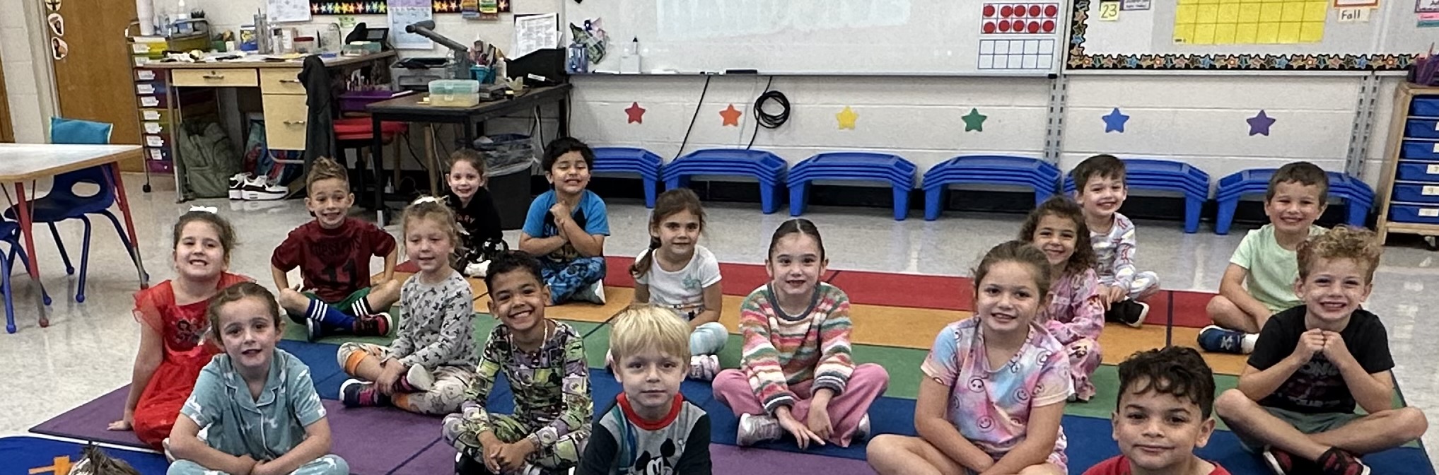 A group of young children in pajamas sit on a colorful mat in a classroom with blue benches and star decorations in the background.