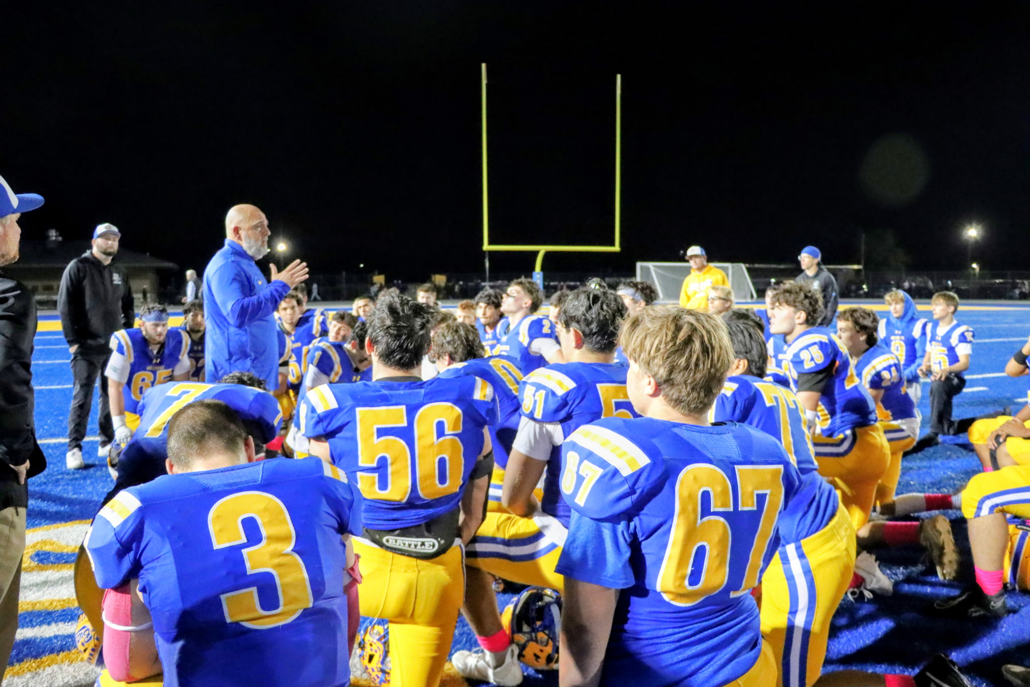 A group of football players in blue and yellow uniforms gathered on a football field at night, with a goal post visible in the background.