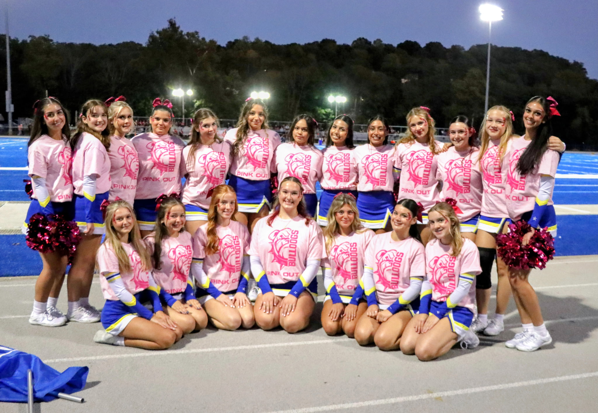 A group of cheerleaders in pink and blue uniforms posing together on a sports field at night, with a brightly lit stadium in the background.