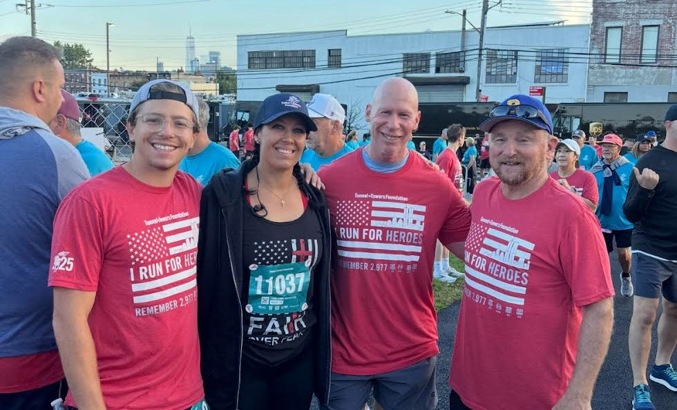A group of people wearing red t-shirts with patriotic designs are gathered together, with a city skyline visible in the background.