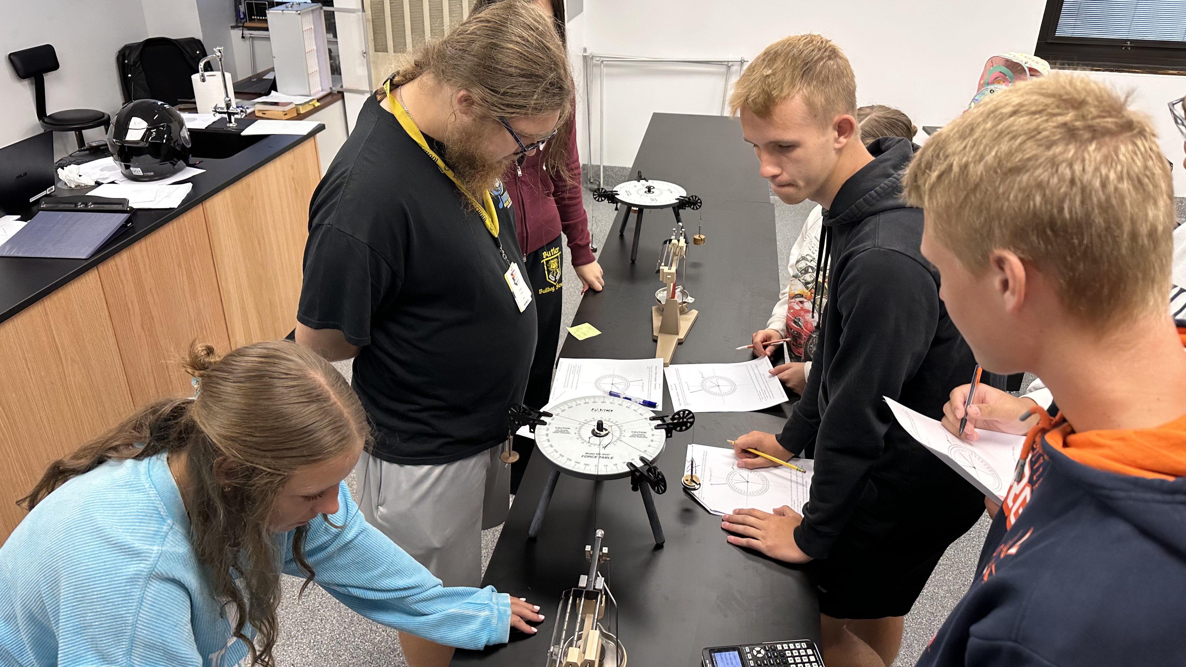 The image shows a group of people gathered around a table, examining what appears to be a piece of equipment or instrument. The background includes various shelves and equipment, suggesting a laboratory or workshop setting.