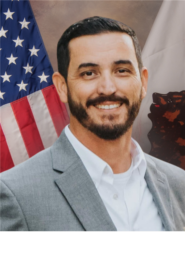 The image shows a smiling man with a beard wearing a gray suit and standing in front of an American flag.