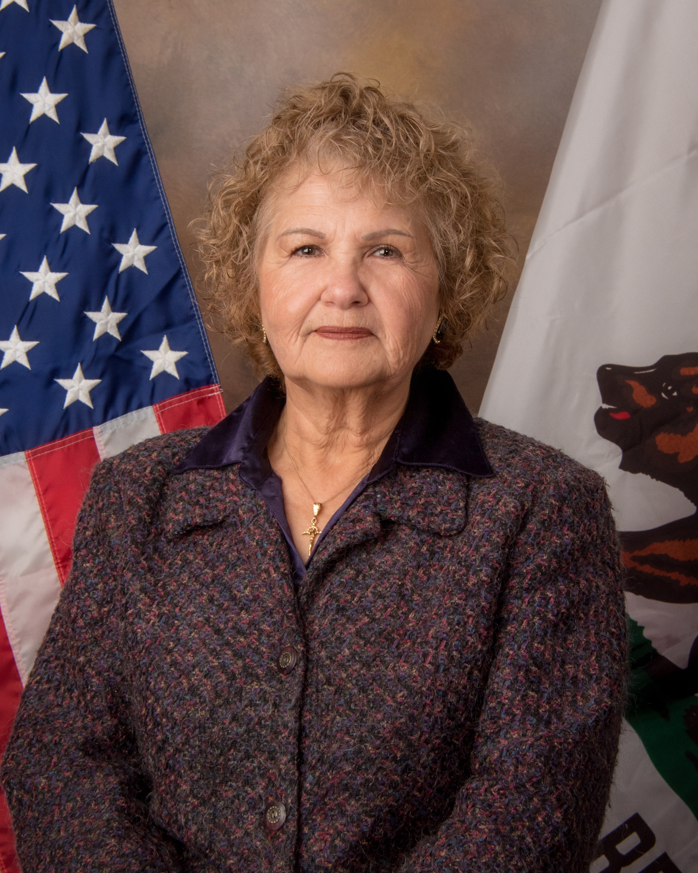 The image shows an elderly woman with curly gray hair wearing a purple and brown patterned jacket, standing in front of an American flag and what appears to be a Native American-style rug or tapestry.