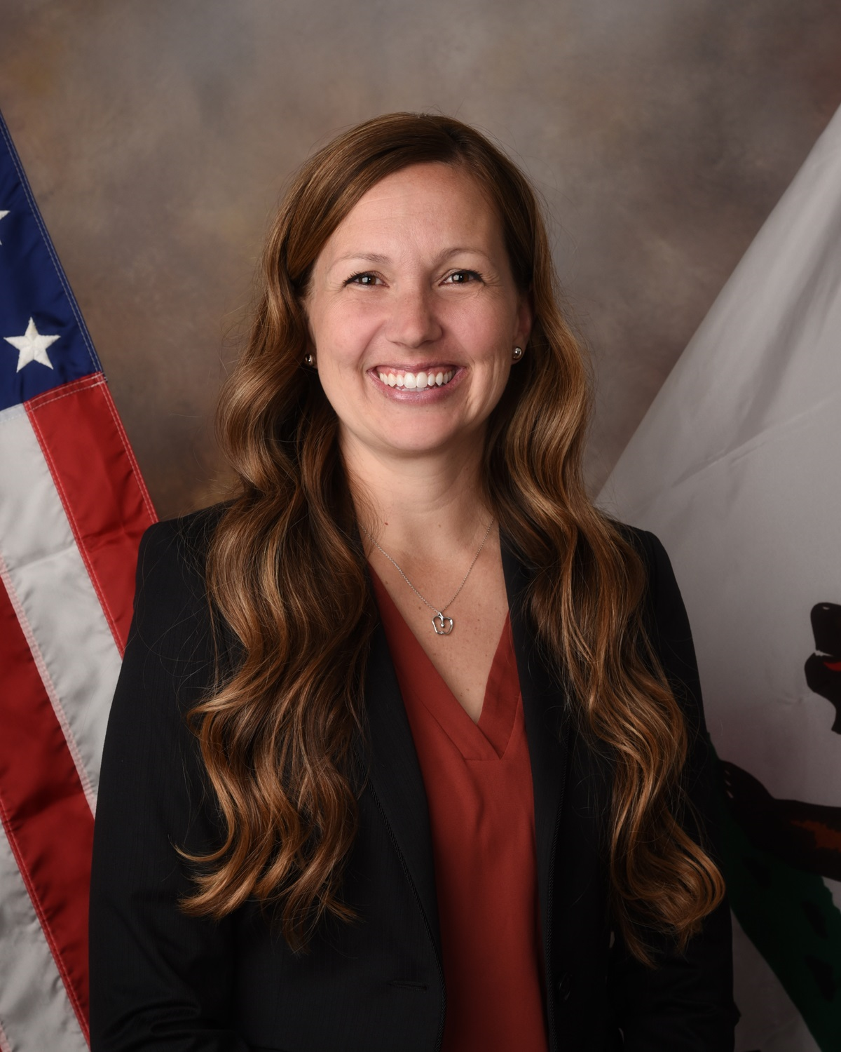 A smiling woman with long, wavy brown hair stands in front of an American flag, wearing a black blazer and red blouse.