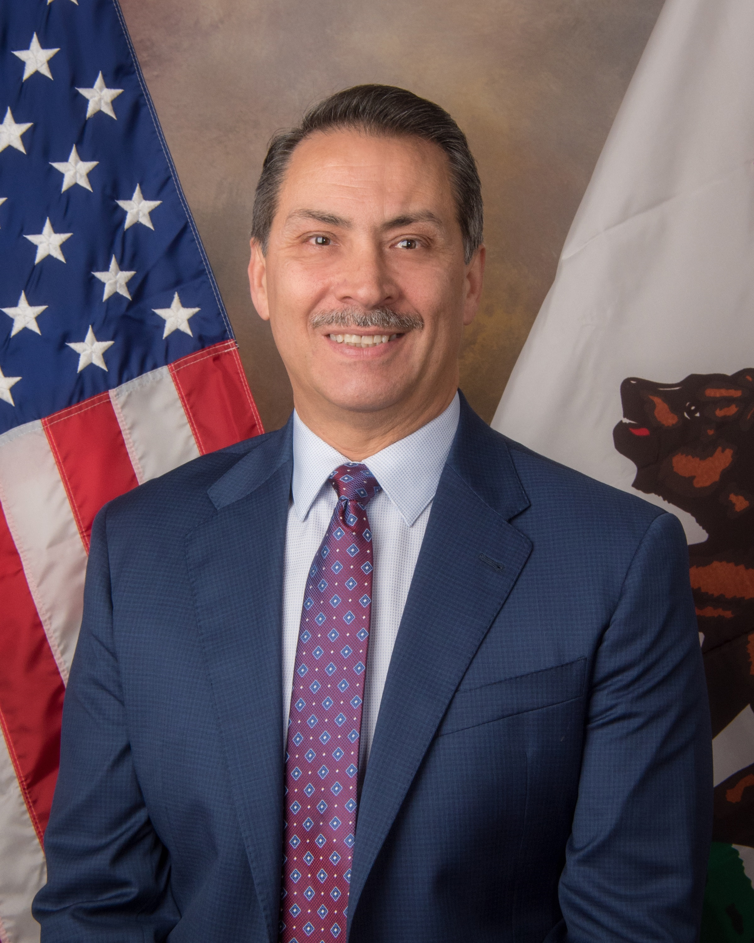 The image depicts a smiling man wearing a navy blue suit and a patterned red tie, standing in front of an American flag.