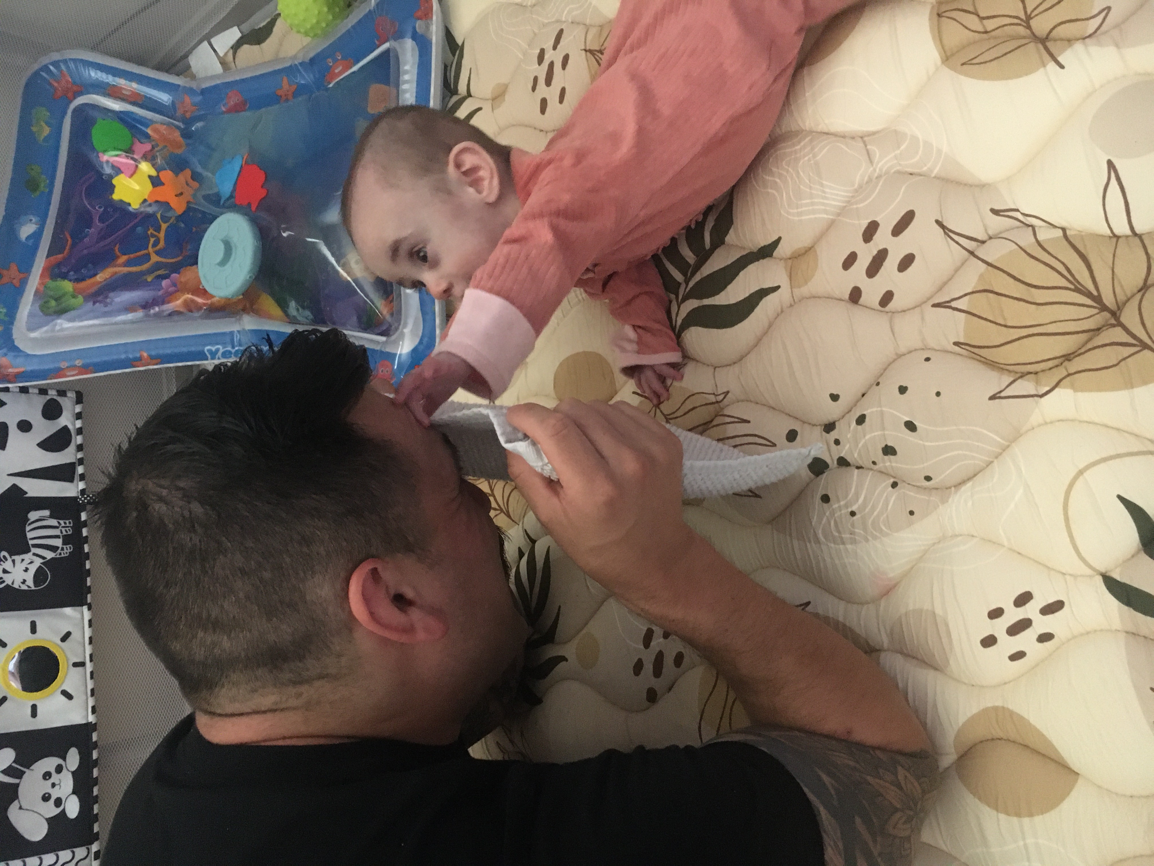 A father and baby are playing on a patterned mat, with a colorful water play mat and black and white animal prints in the background.