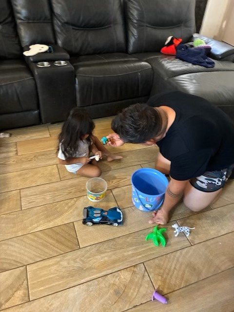 A man and a young child play with toys on a wooden floor in front of a large black leather couch.