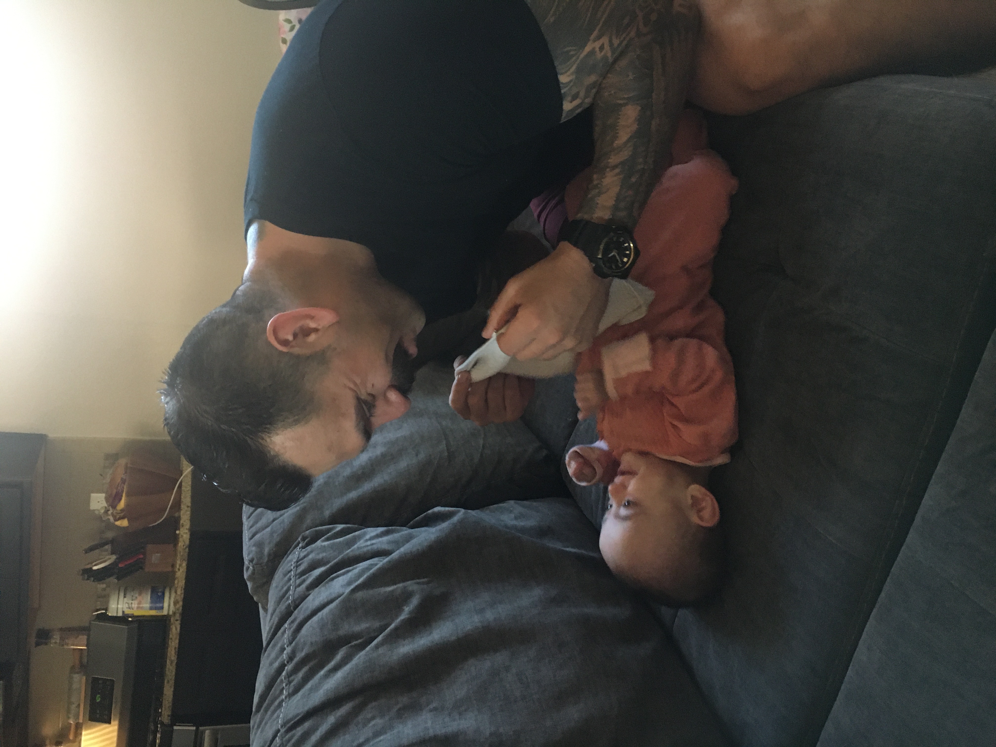 A man in a black t-shirt plays with a baby in a pink outfit on a gray couch, with a blurred kitchen counter in the background.