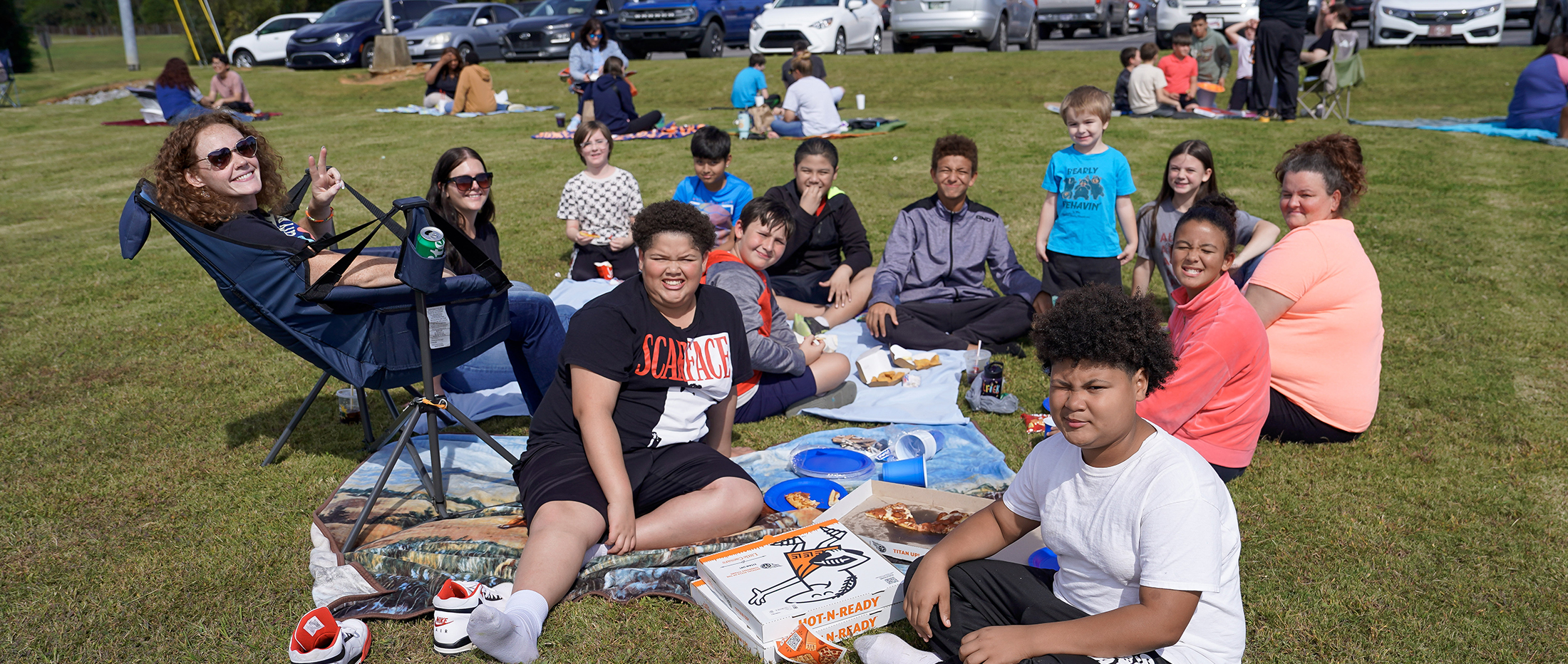 students and teachers seated on a green field on blankets eating lunch