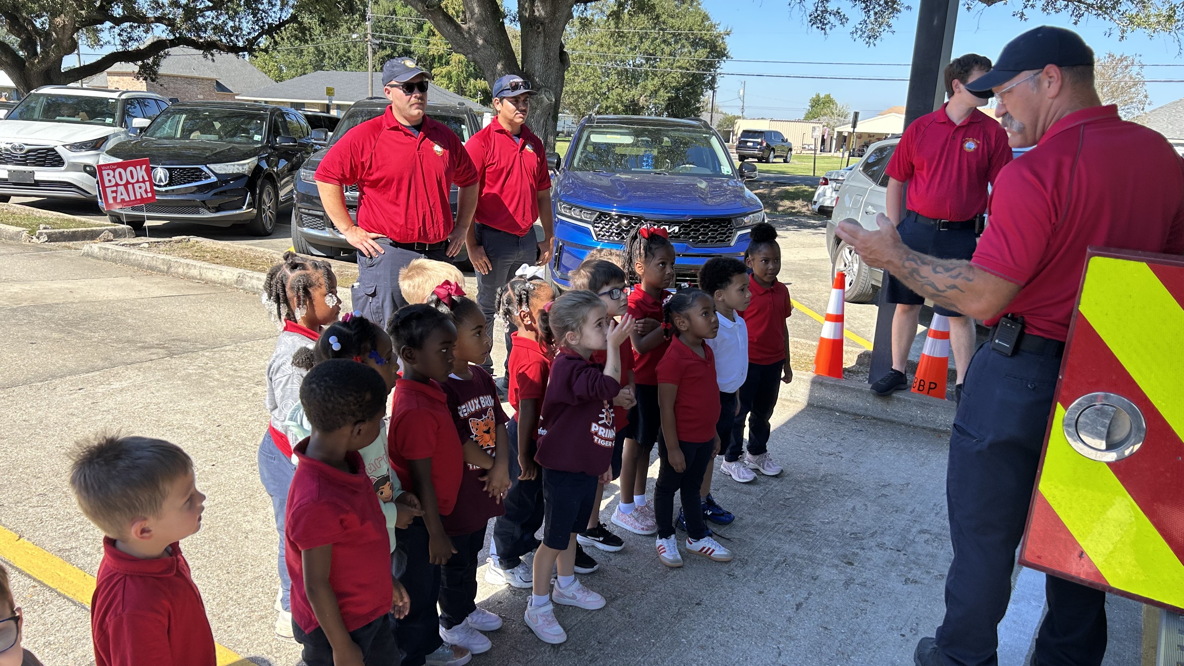 A group of firefighters in red shirts interact with young children in red shirts, with cars and trees visible in the background.