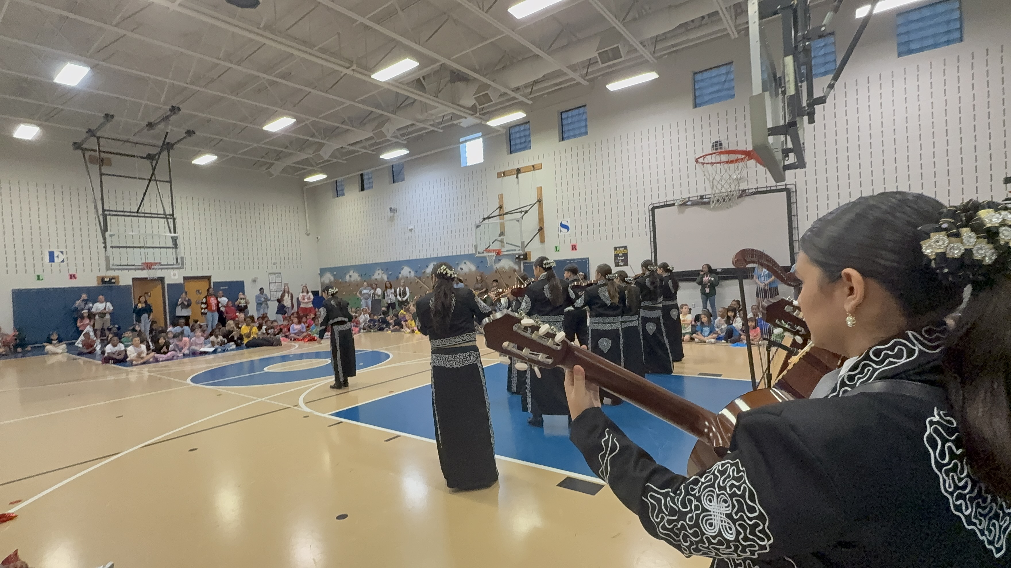 A large indoor gymnasium with a basketball court, filled with people playing instruments and performing on stage.