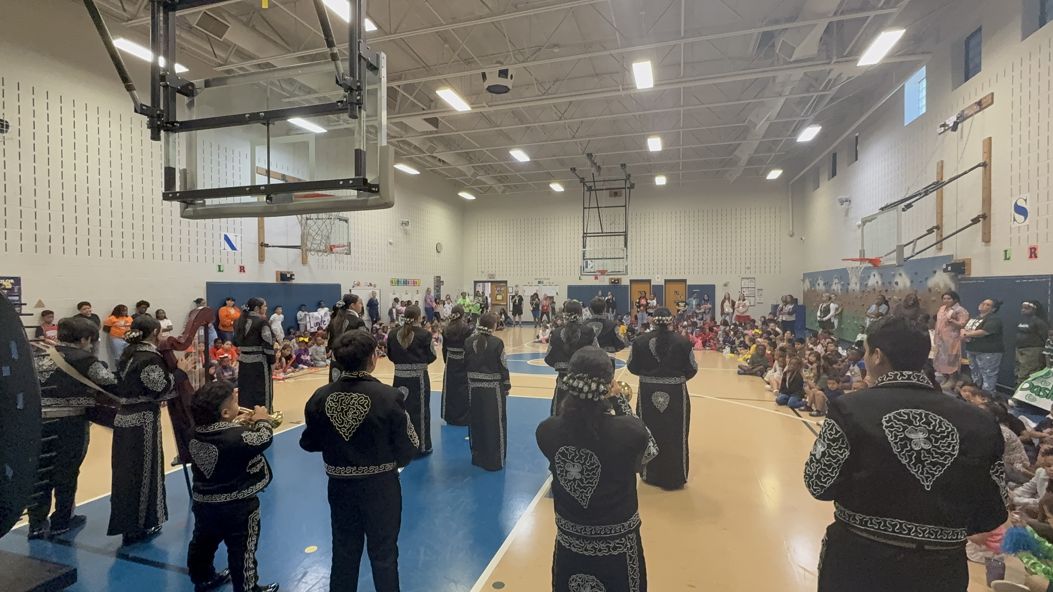 A crowded indoor gymnasium filled with people wearing dark clothing and standing in rows, with basketball hoops and other sports equipment visible in the background.