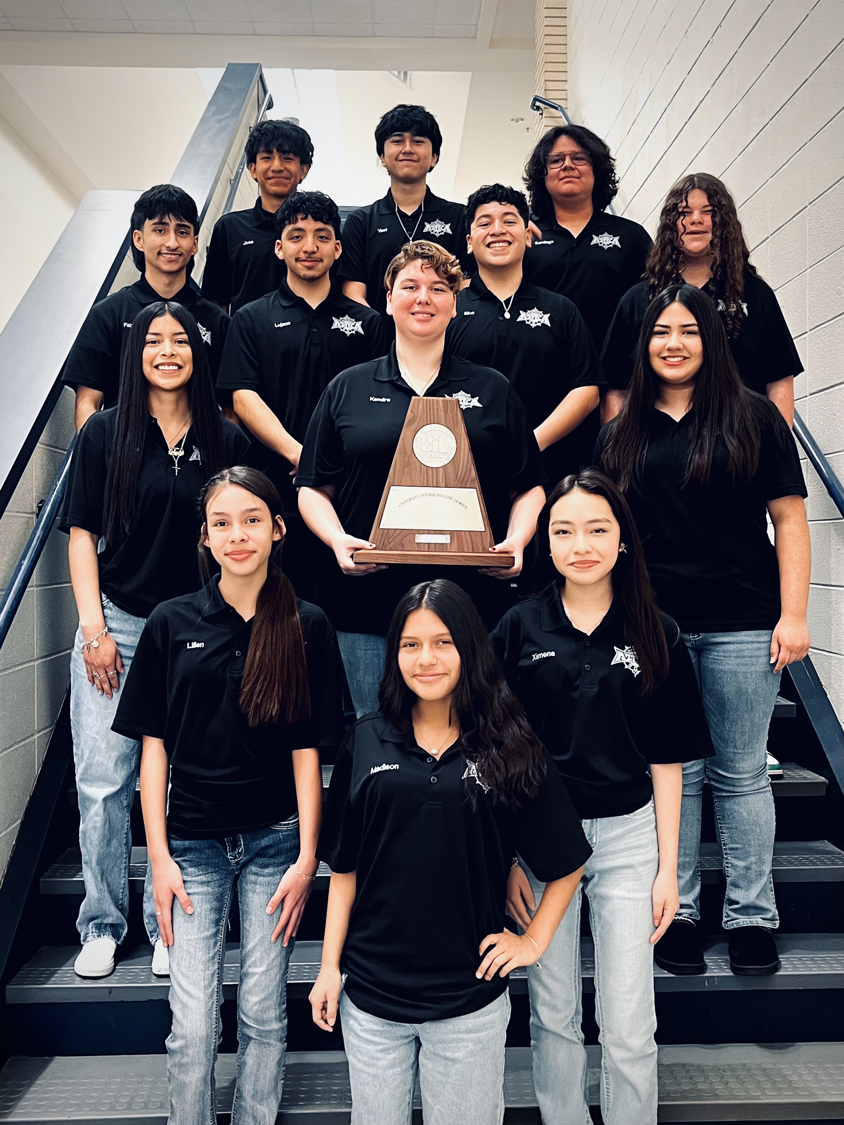 A group of young people, likely a sports team or club, posing together with a trophy or award in the foreground. They are wearing matching black shirts and appear to be in a school or indoor setting.