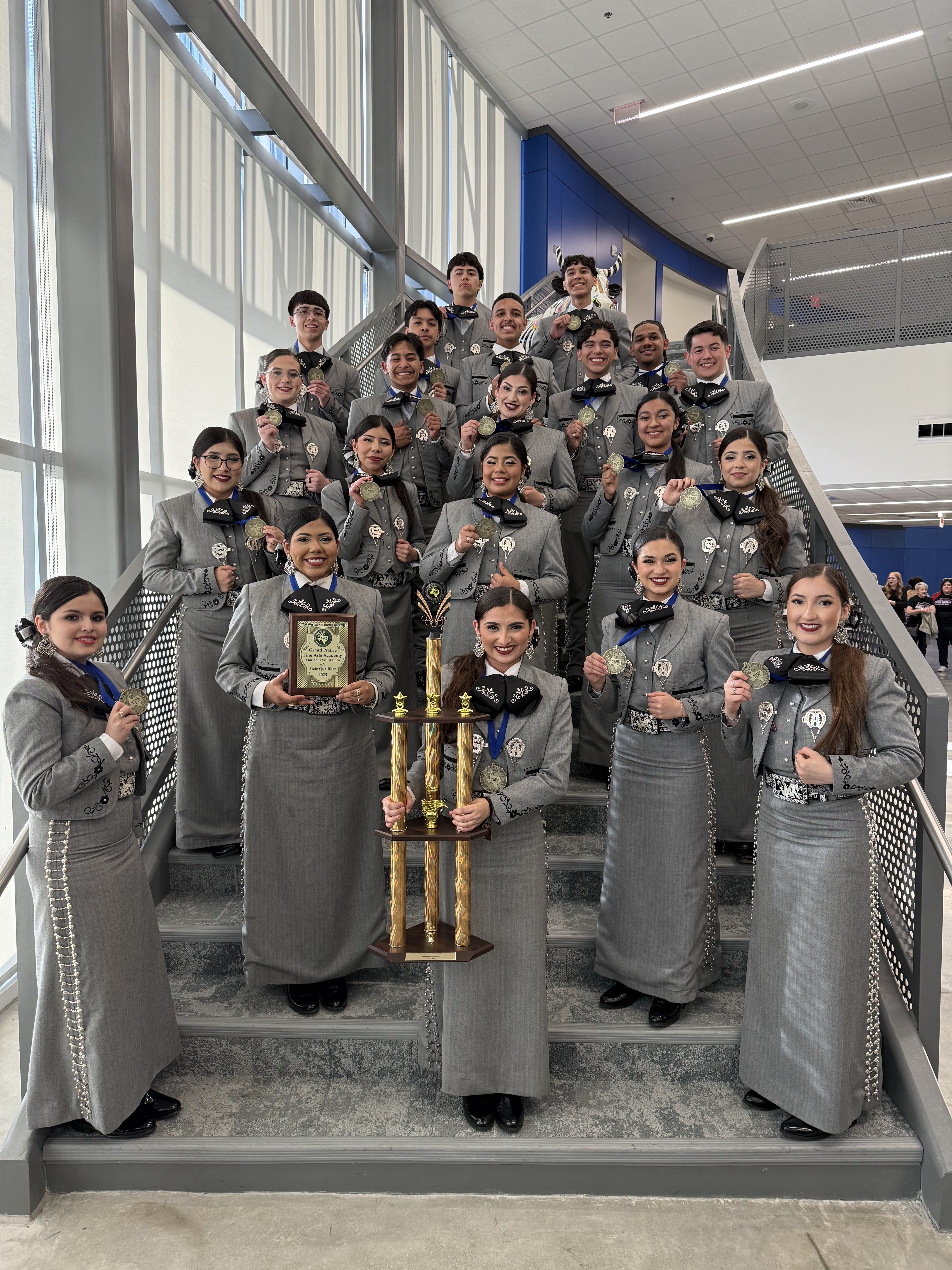 A group of young people, likely students, dressed in gray uniforms are gathered on a staircase, holding trophies and posing for a photograph.