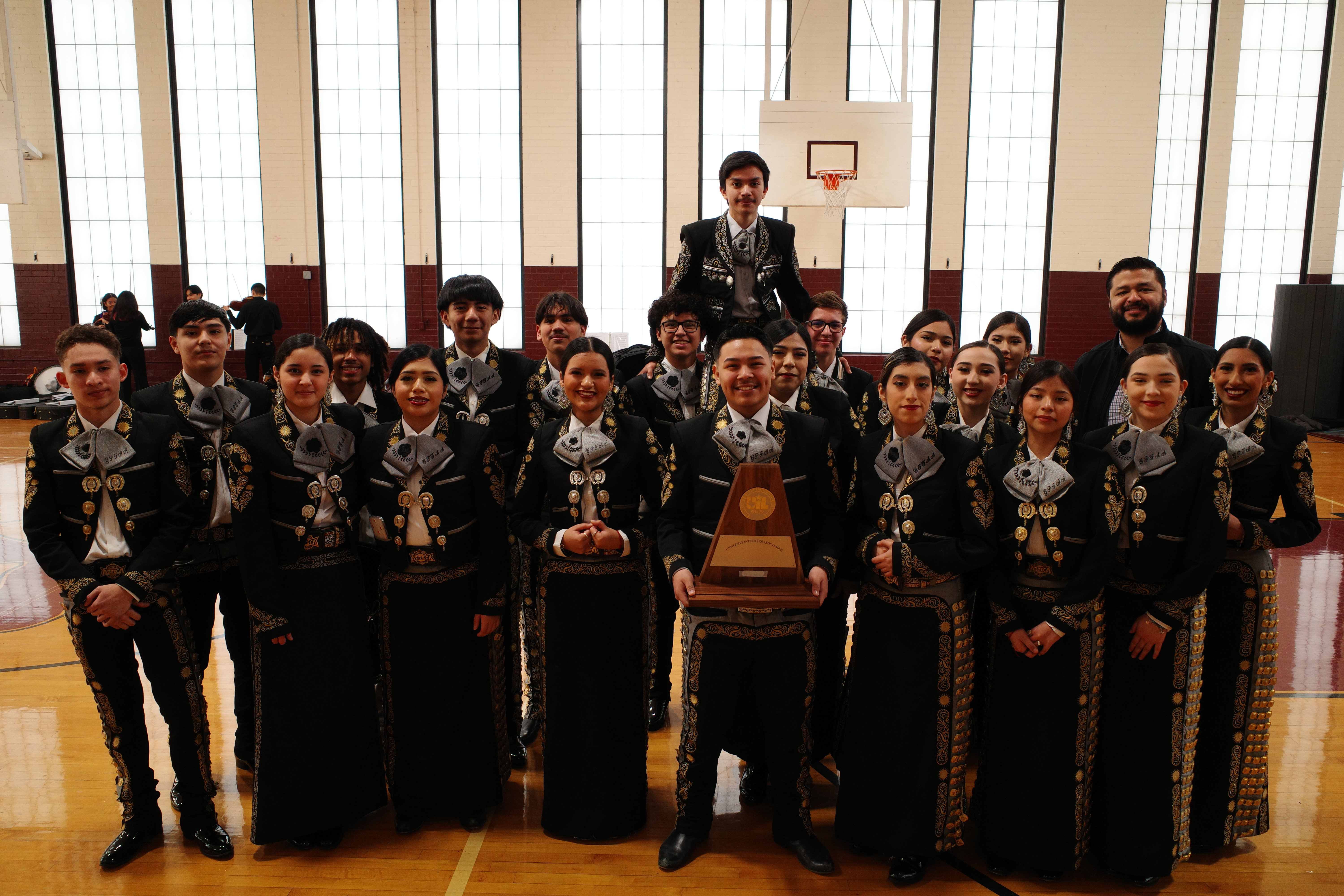 A large group of people dressed in traditional Mongolian clothing, standing together in a formal setting with a basketball hoop visible in the background.