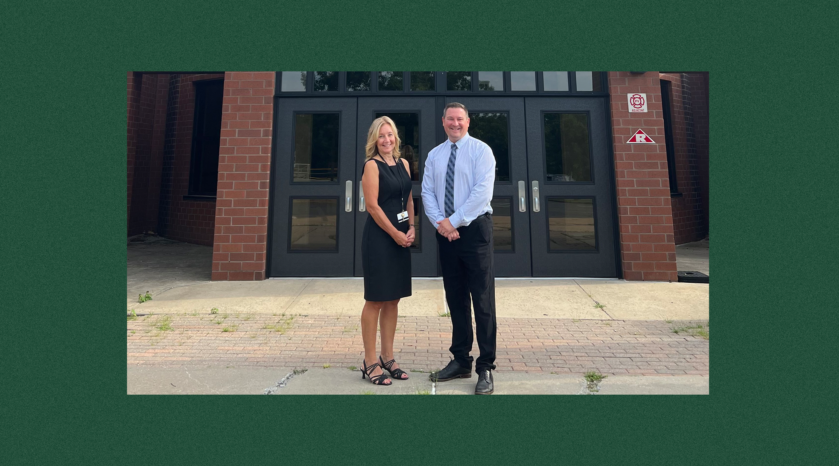 A man and a woman are standing together in front of a brick building, with the woman wearing a black dress and the man wearing a light blue shirt.