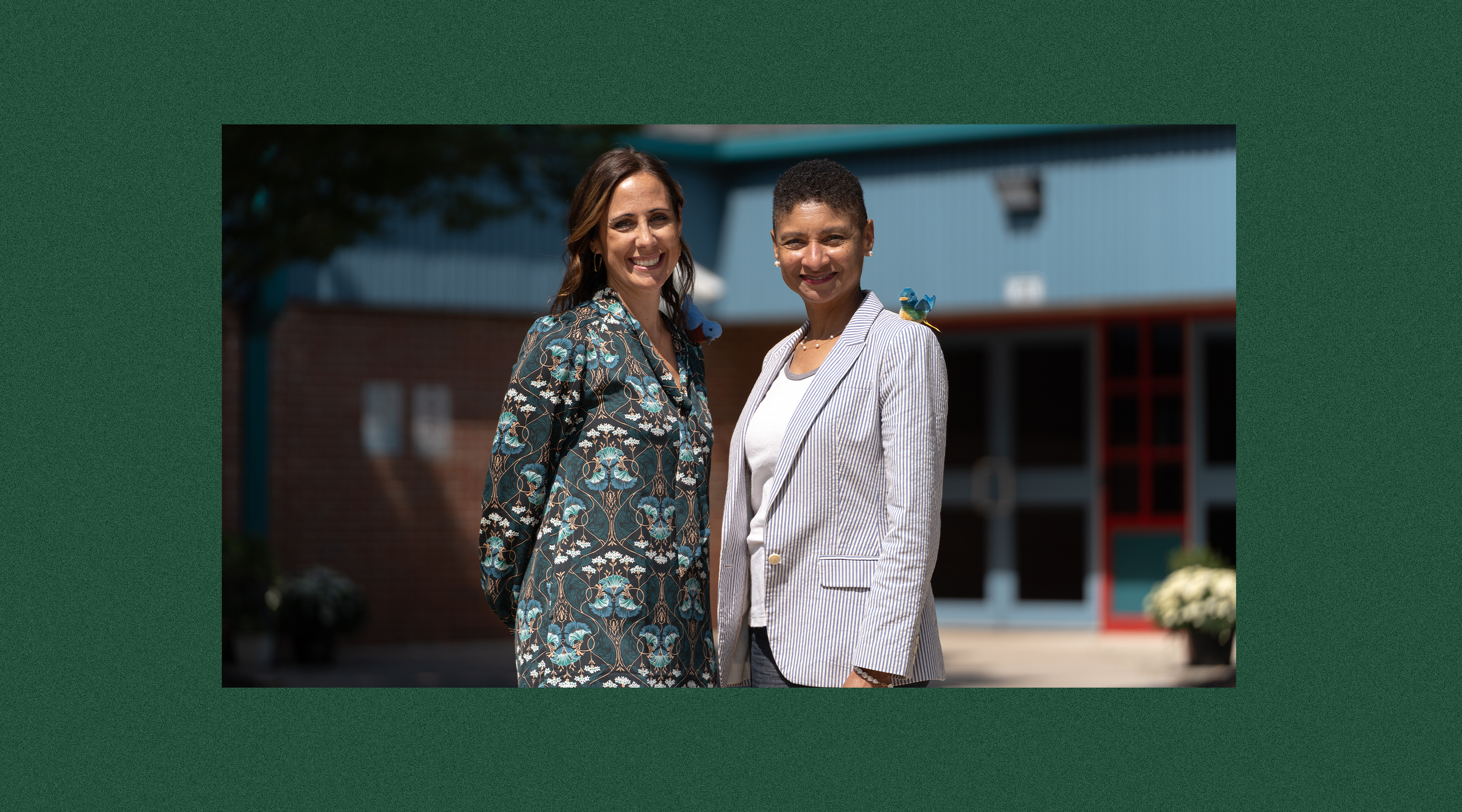 Two women standing together in front of a building, one wearing a floral dress and the other a white blazer.
