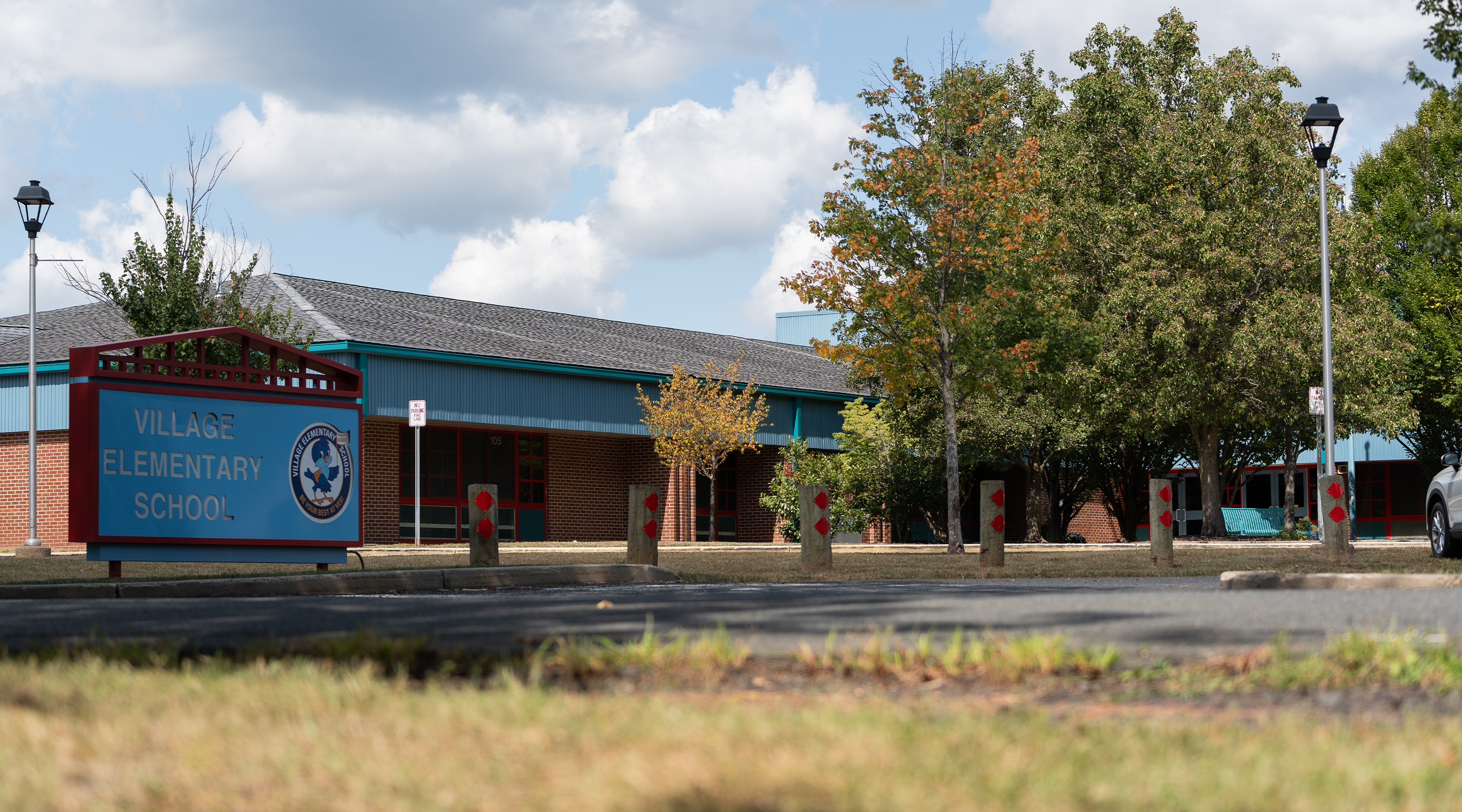 A brick elementary school building with a "Village Elementary School" sign, surrounded by trees and a grassy area in the foreground, under a partly cloudy sky.