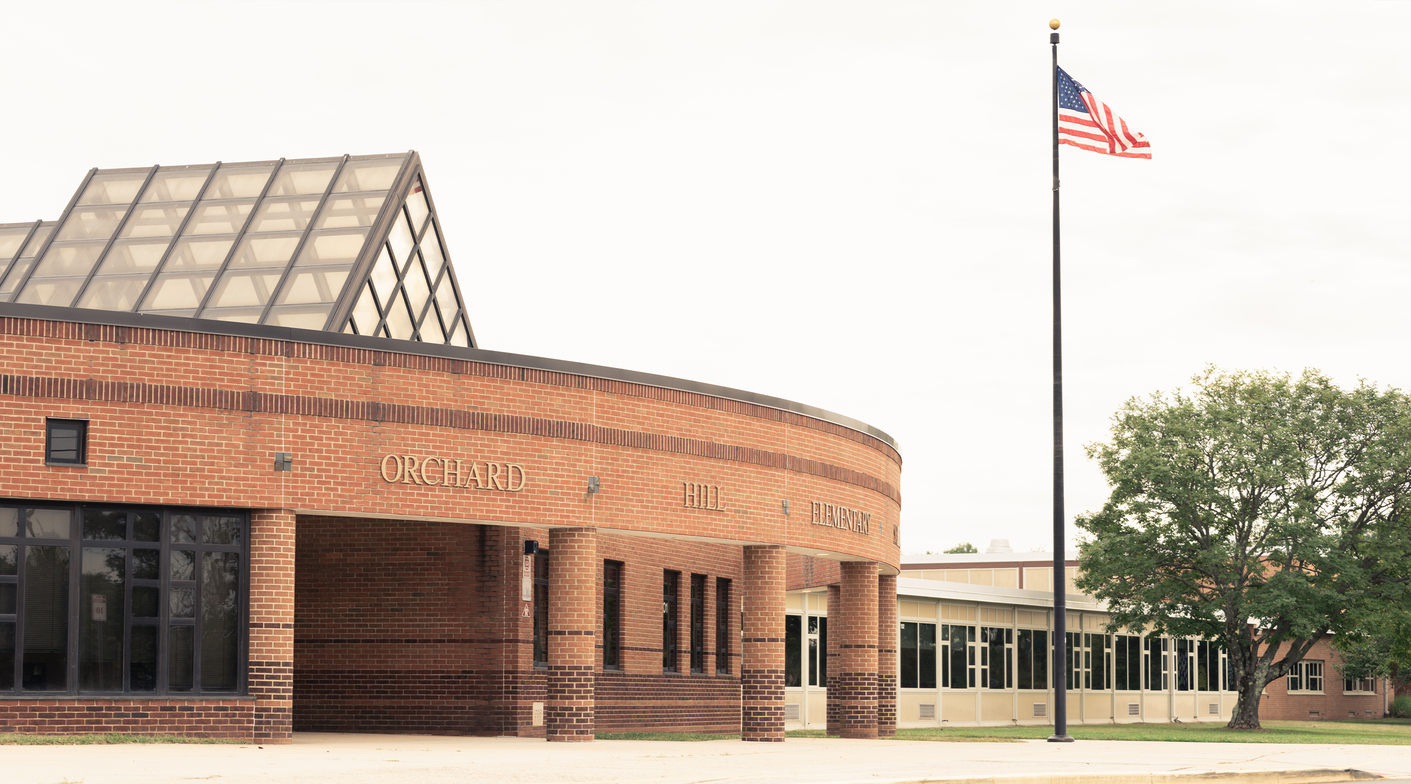 A brick building with a curved roof and large windows, with an American flag flying in the foreground and trees in the background.