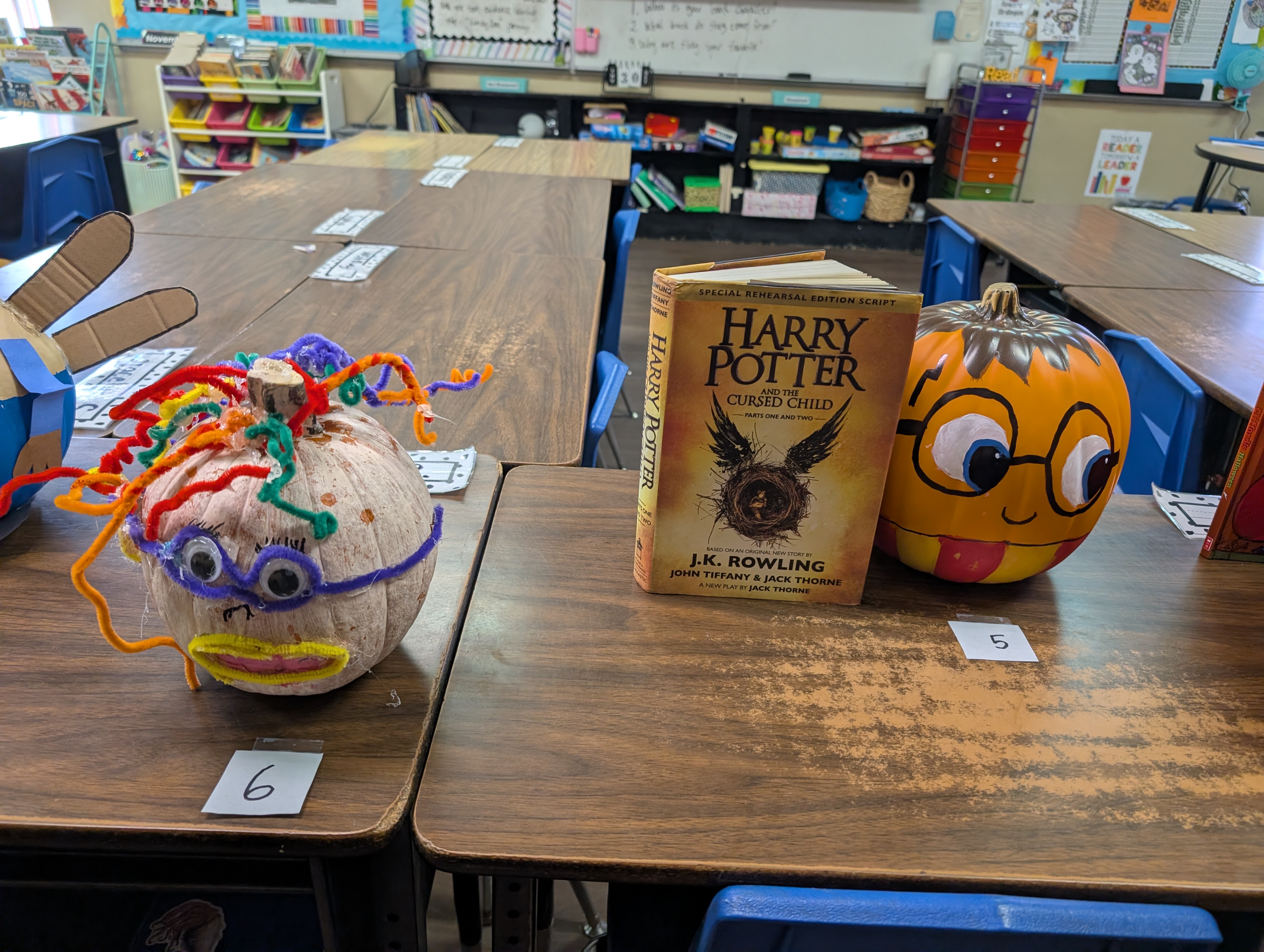 The image shows a classroom setting with various educational materials and decorations, including a Harry Potter book, a pumpkin-shaped decoration, and colorful craft projects on a desk.