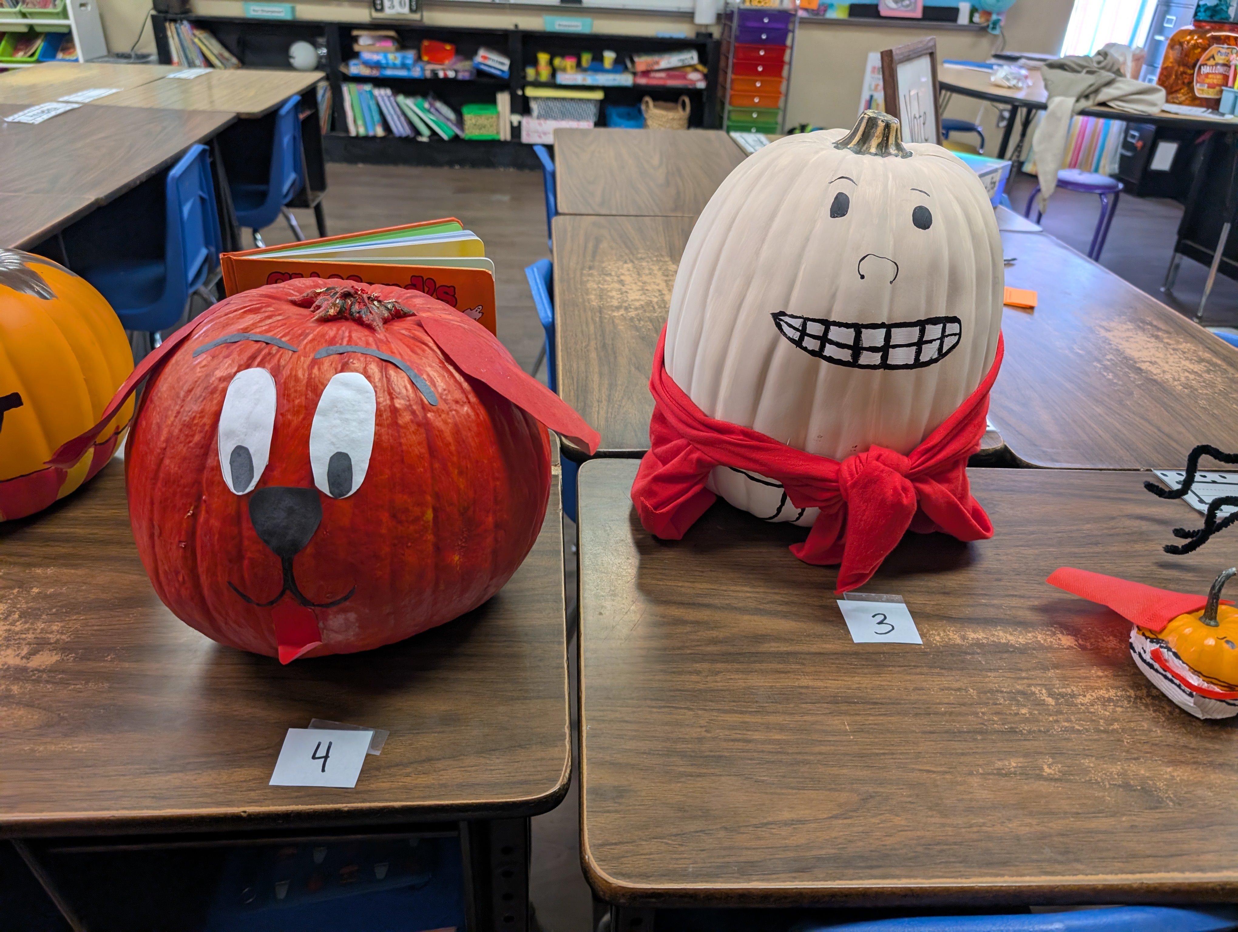 The image shows two decorated pumpkins on a classroom desk, with various school supplies and materials visible in the background.