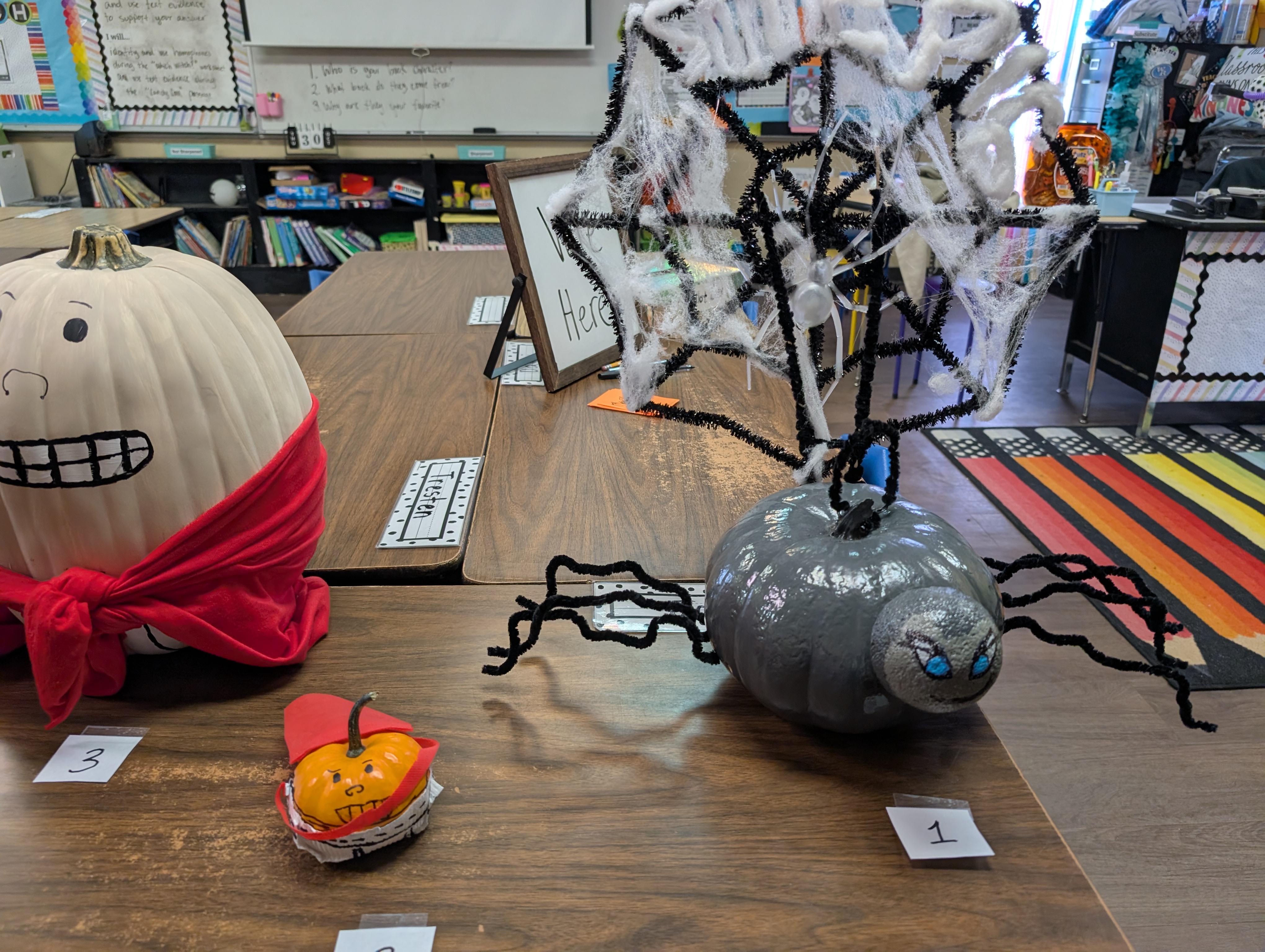 The image shows a classroom or educational setting with various Halloween-themed decorations and props, including a large pumpkin, a spider-like creature, and other spooky elements, all arranged on a wooden floor.