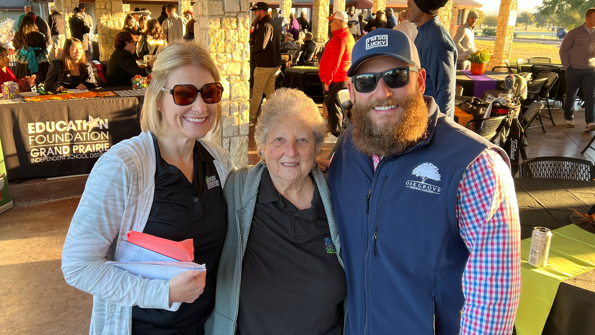 The image shows three people standing together in what appears to be an outdoor market or fair setting, with various clothing and other items displayed in the background.