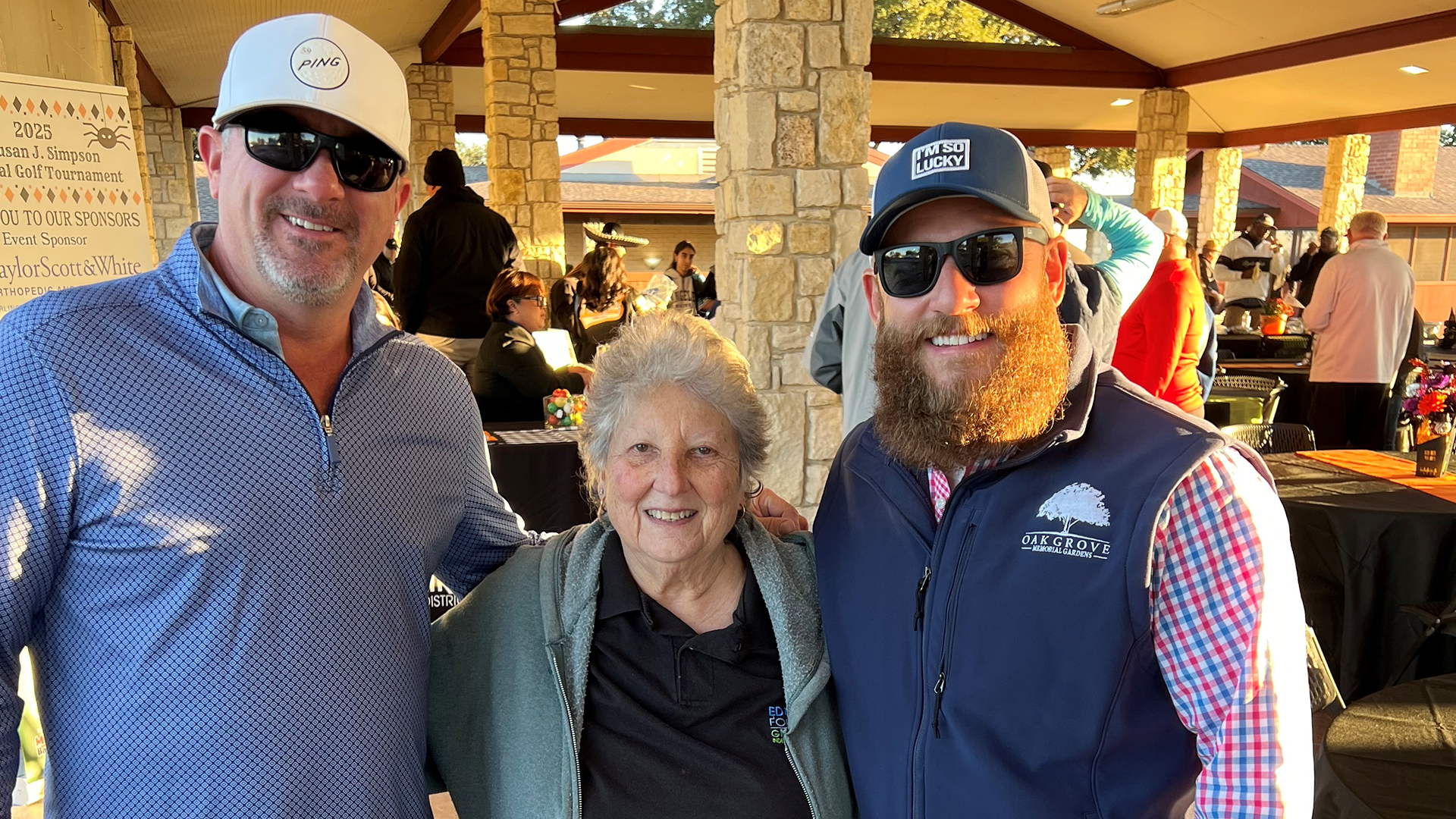 The image shows three people standing together, with an older woman in the center flanked by two men, one with a beard and the other with a baseball cap.