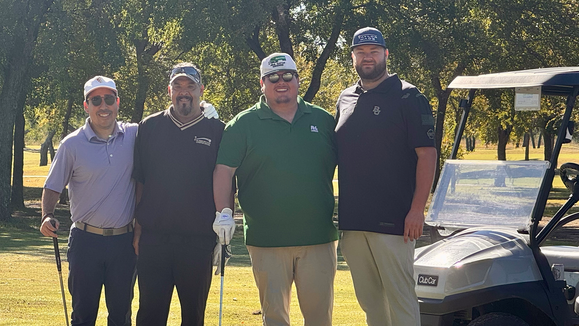 The image shows four men standing together on a golf course, with a golf cart visible in the background.