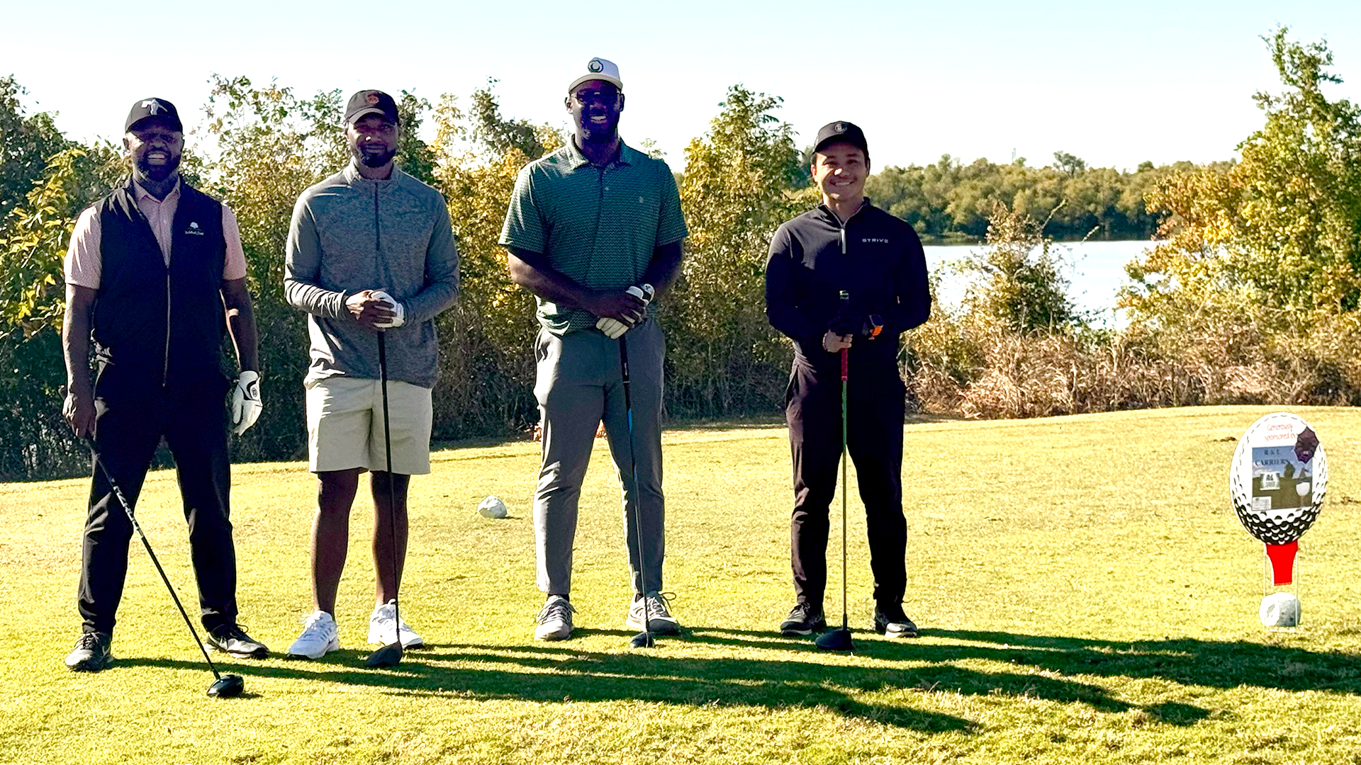 A group of people dressed in athletic attire standing on a grassy field with trees and a clear sky in the background.