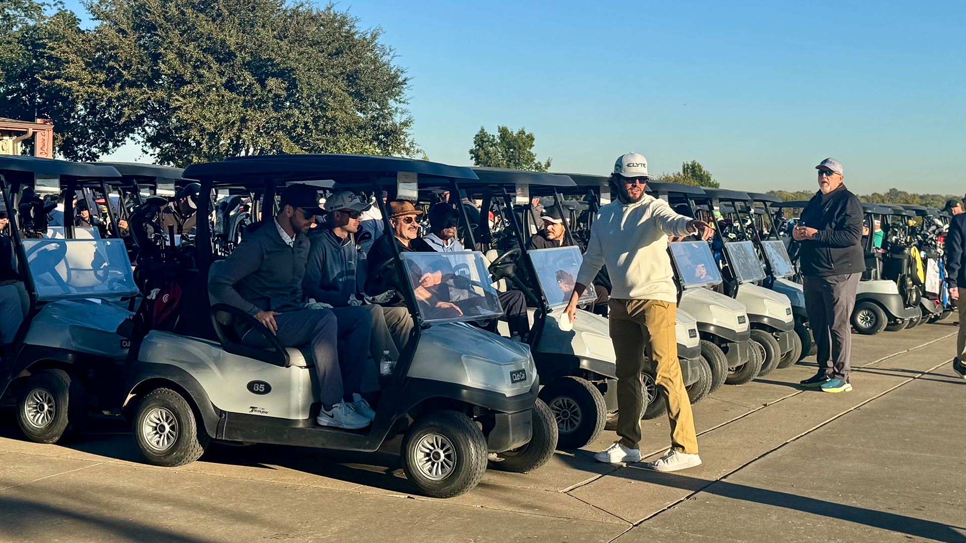 A group of people standing next to several golf carts in an outdoor setting with trees and a clear sky in the background.
