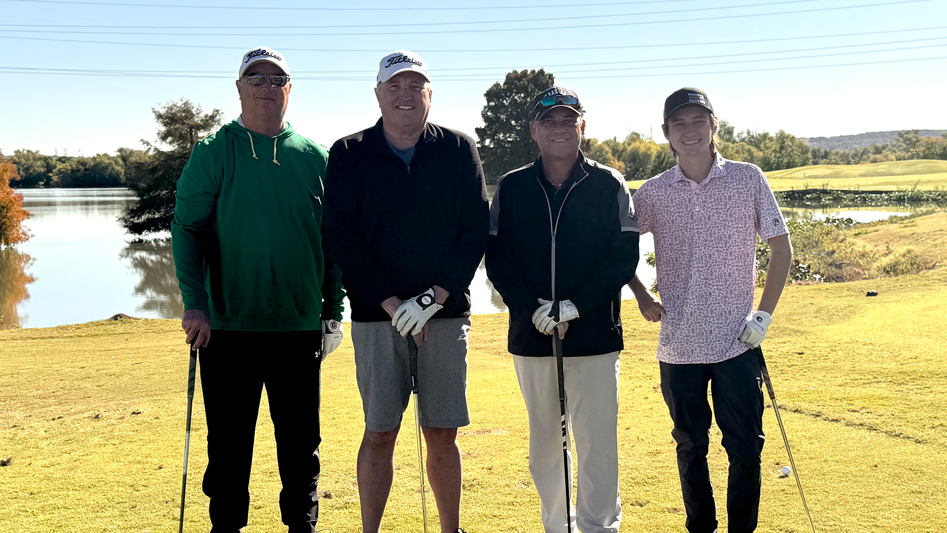 Four individuals, dressed in casual attire, stand together on a golf course with a lake visible in the background.