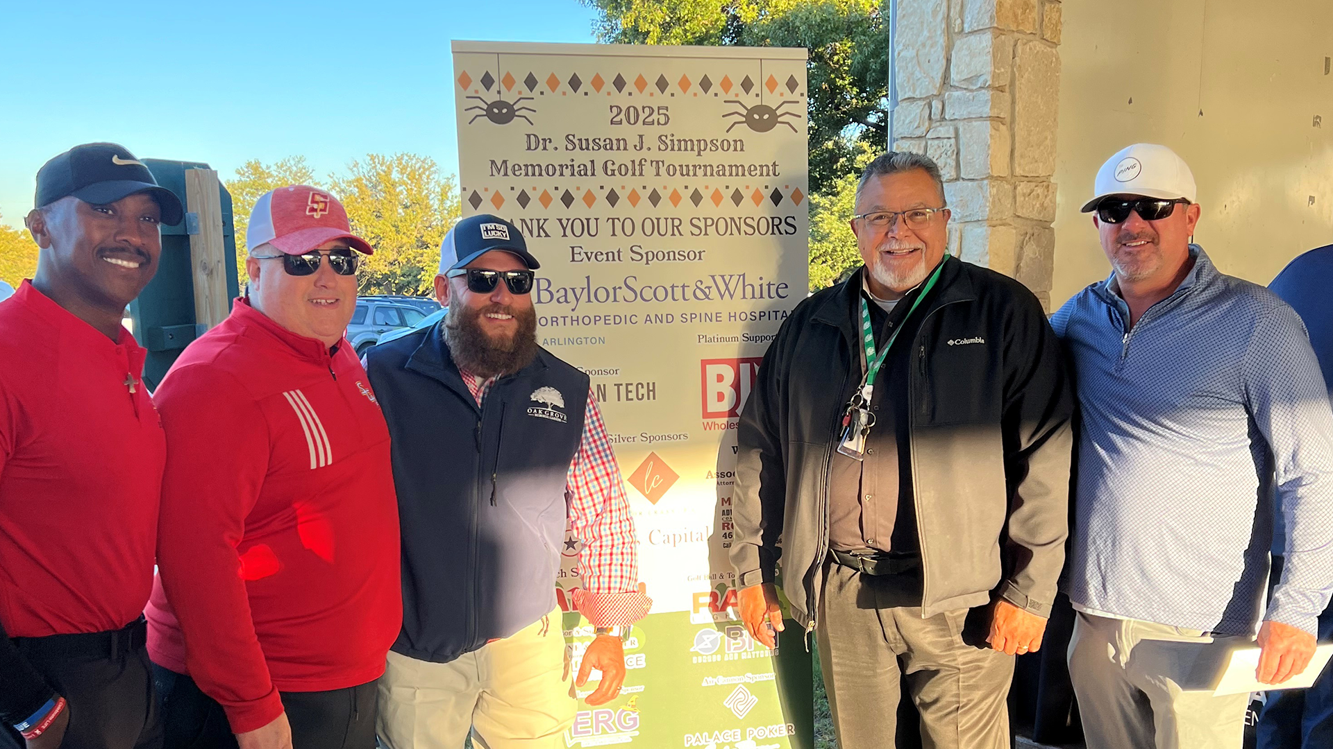 A group of men, some wearing casual clothing and others in more formal attire, stand in front of a sign displaying the name "Dr. Susan J. Simpson Memorial Golf Tournament" against a backdrop of trees and a clear blue sky.