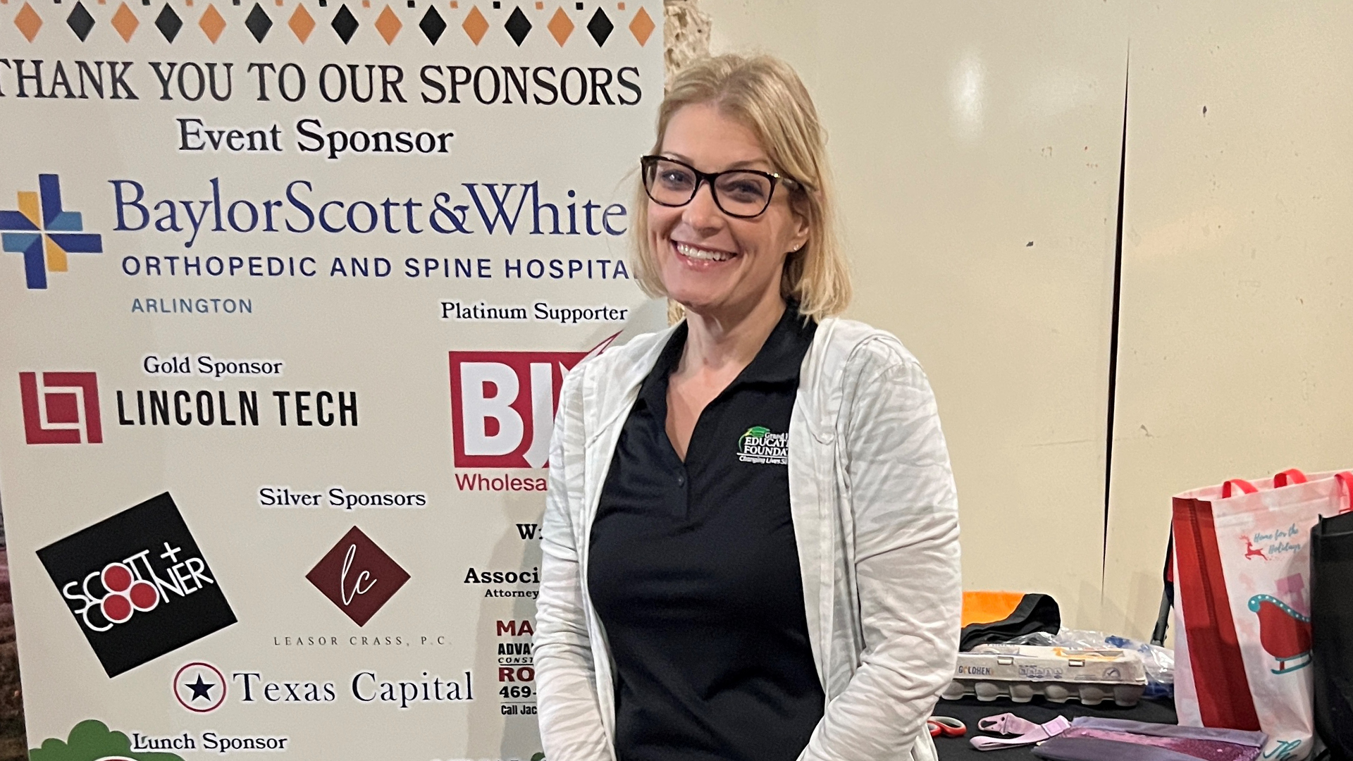 The image shows a woman wearing glasses and a white jacket standing in front of a display with the names of various sponsors for an event.