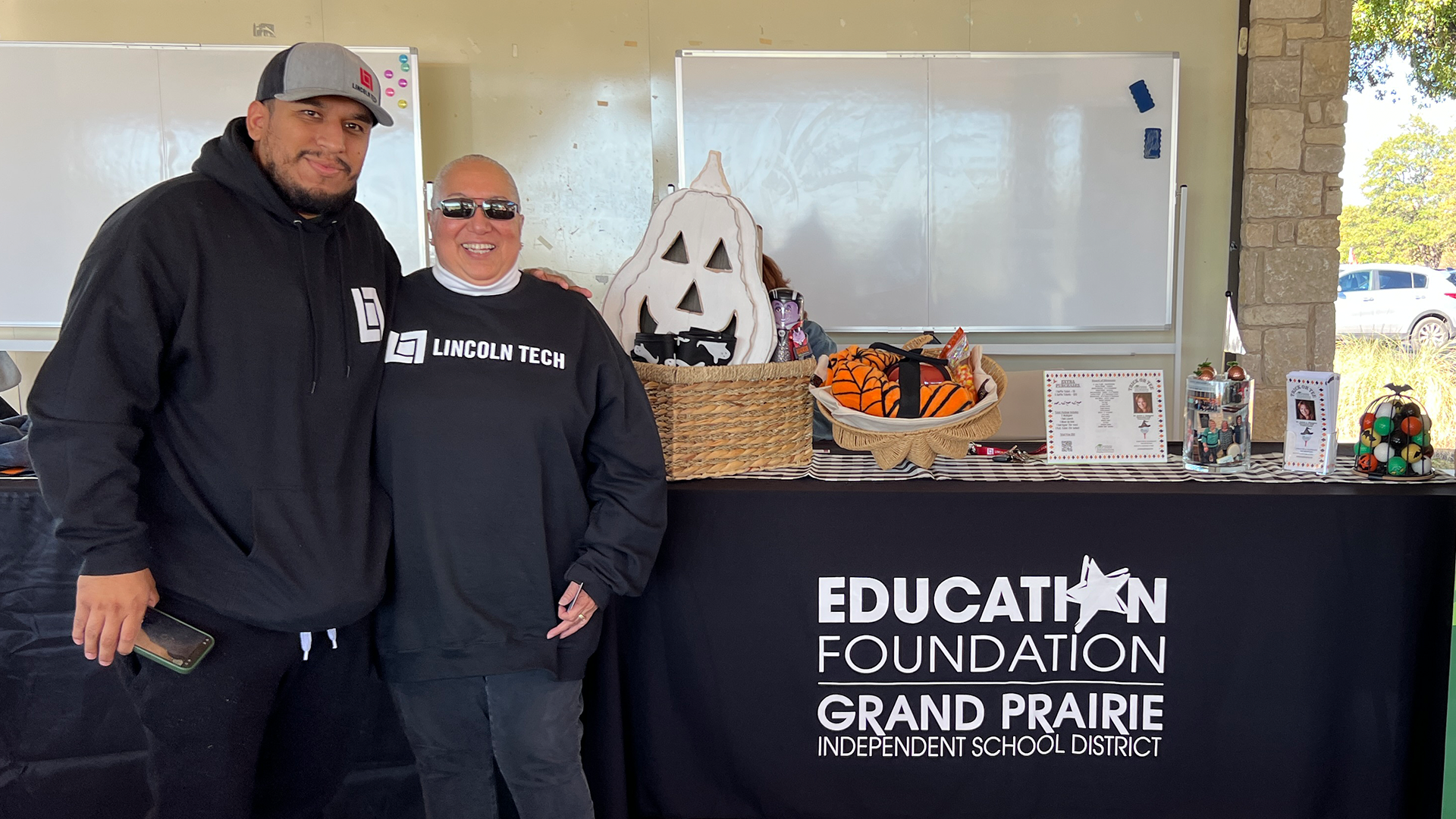 Two individuals wearing hoodies and hats are standing in front of a table with various items, including a basket and a whiteboard displaying the text "Education Foundation Grand Prairie".