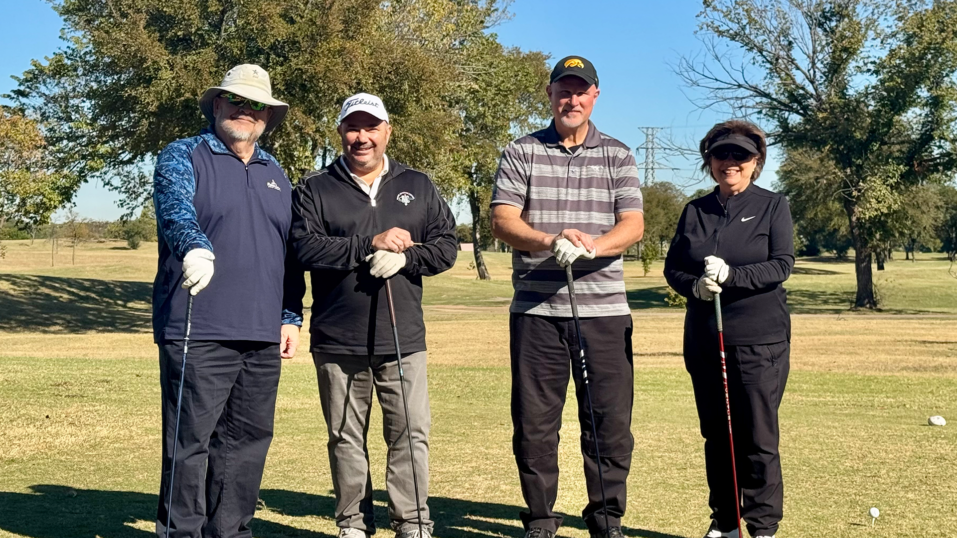 Four people, dressed in golf attire, standing together on a grassy golf course with trees in the background.