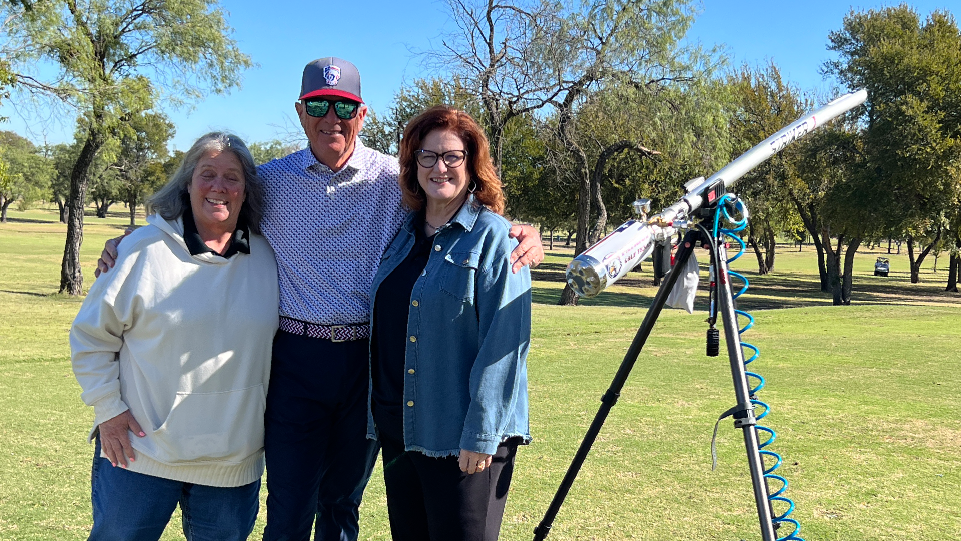 The image shows three people standing together in a park-like setting, with trees and a clear blue sky in the background.