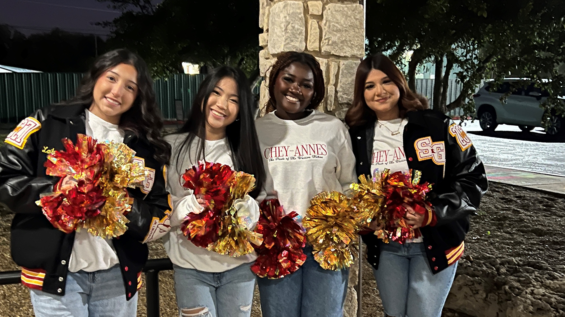 Four young women in casual clothing are standing together, holding colorful pom-poms, in front of a stone structure at night.