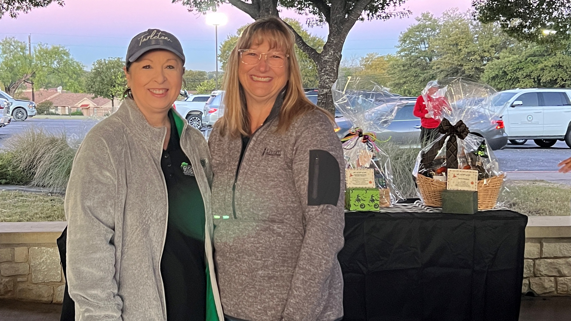 Two women wearing casual clothing and coats are standing together in an outdoor setting, with a table in the background displaying various items.