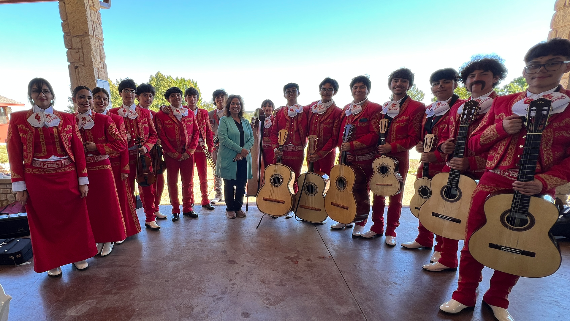 A group of people wearing traditional red costumes and holding guitars stand in front of a wooden structure against a clear blue sky.
