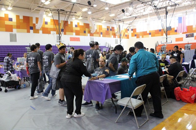 In the foreground, people are donating blood at tables, while the background shows a gymnasium with bleachers and flags.